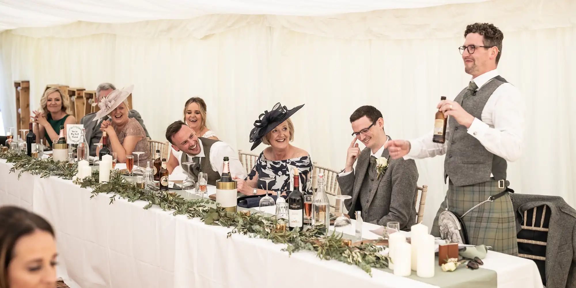 A group of people, dressed formally, are seated at a long, decorated table. One man stands, speaking with a bottle in hand, while the rest laugh and listen attentively.