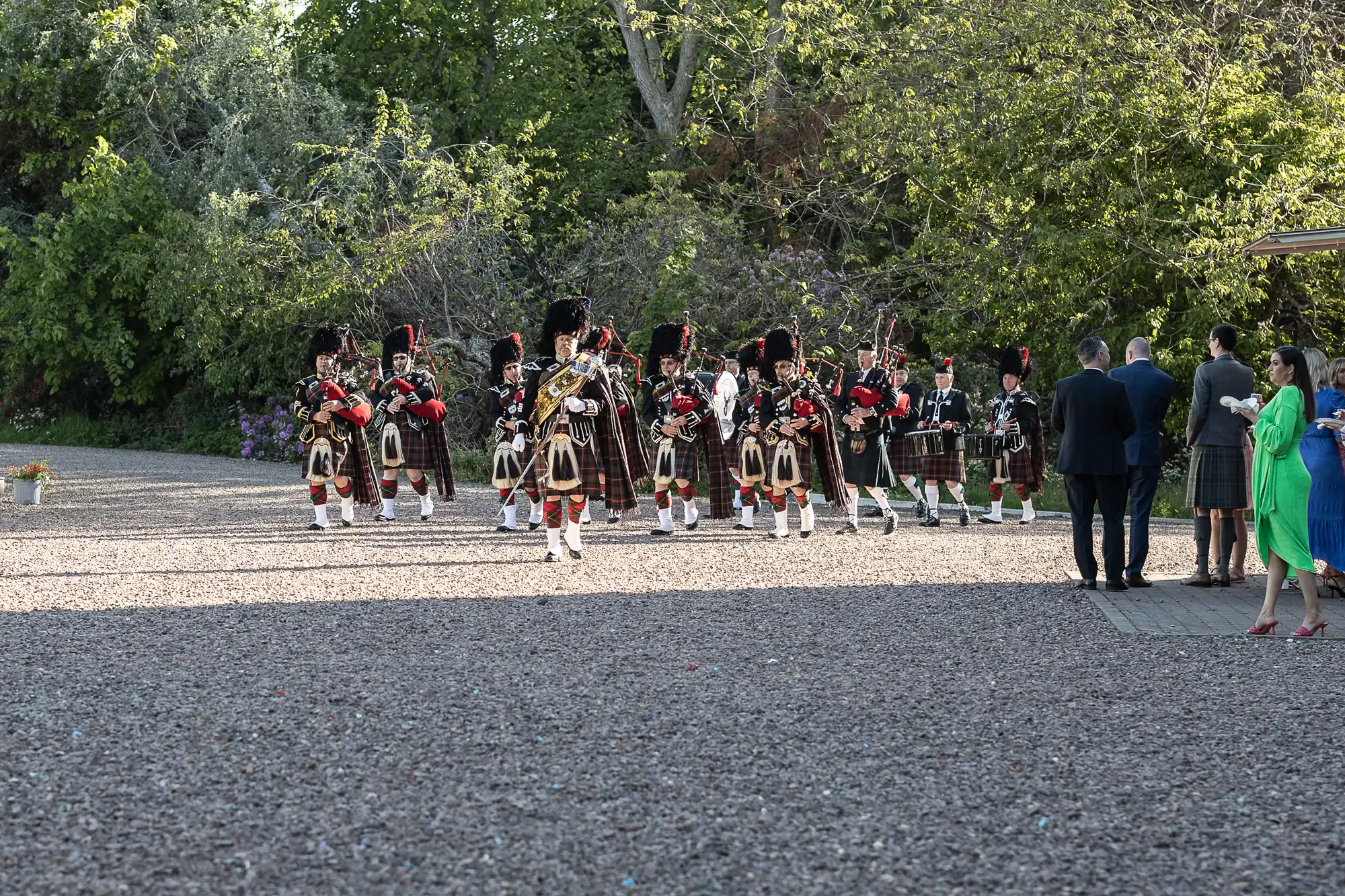 A group of people in formal attire watches a pipe band dressed in traditional Scottish kilts perform outdoors on a gravel area with trees in the background.