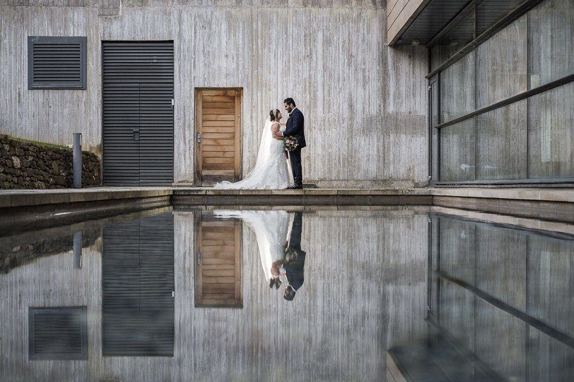Norton House Hotel wedding photography - relaxed and natural couple portraits by Norton House Hotel wedding photographer: A bride and groom stand close together by a wooden door, their reflection visible in a calm, shallow pool in front of a modern building.