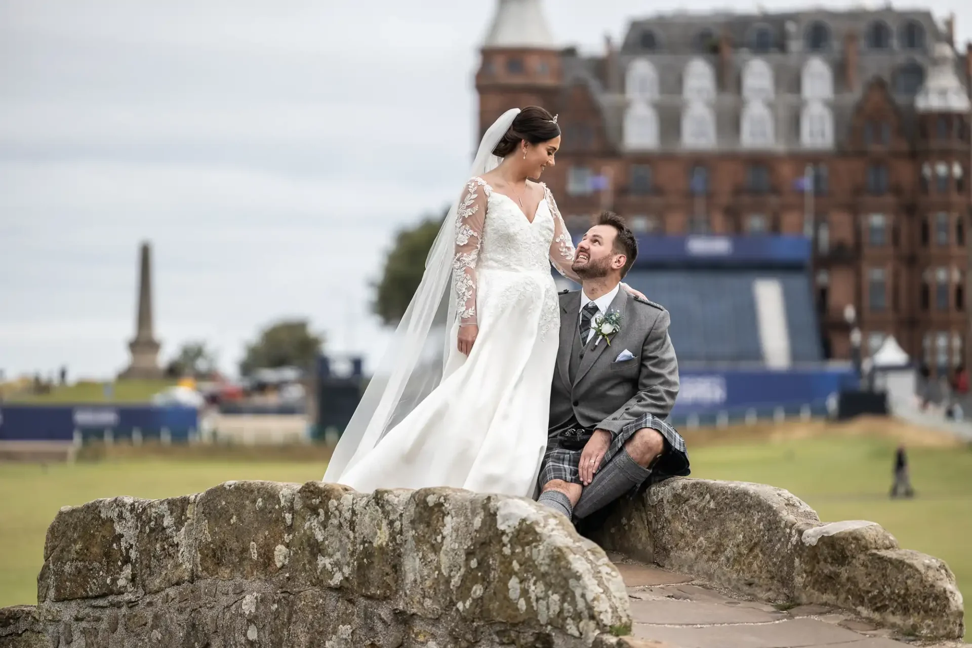 Old Course Hotel wedding photographer: Bride in a white gown and groom in a kilt sitting on a stone bridge, with a historic building and obelisk in the background.