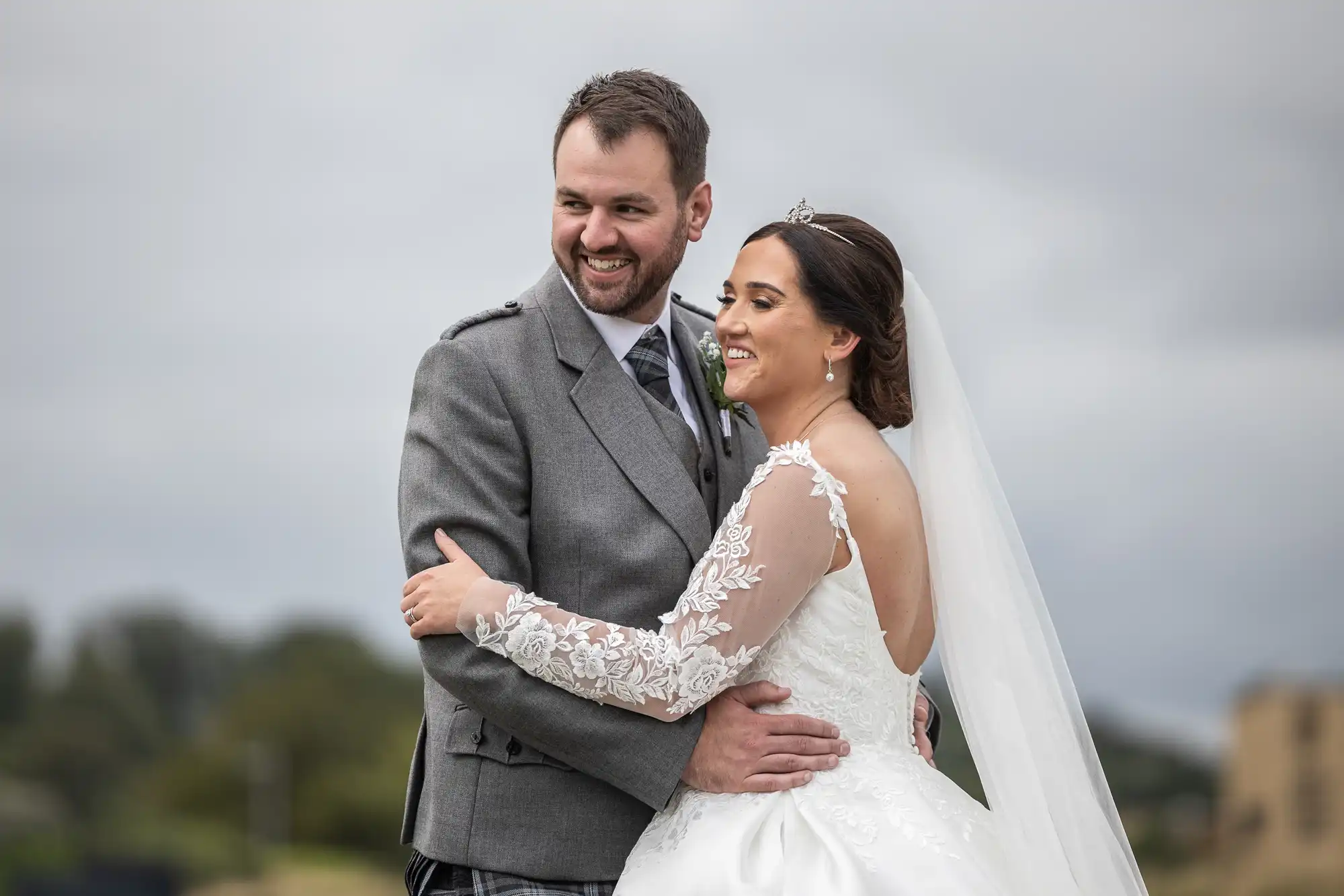 A bride and groom embrace outdoors, both smiling. The bride wears a white gown with lace sleeves and a veil; the groom is in a gray suit with a tie. Overcast sky in the background.