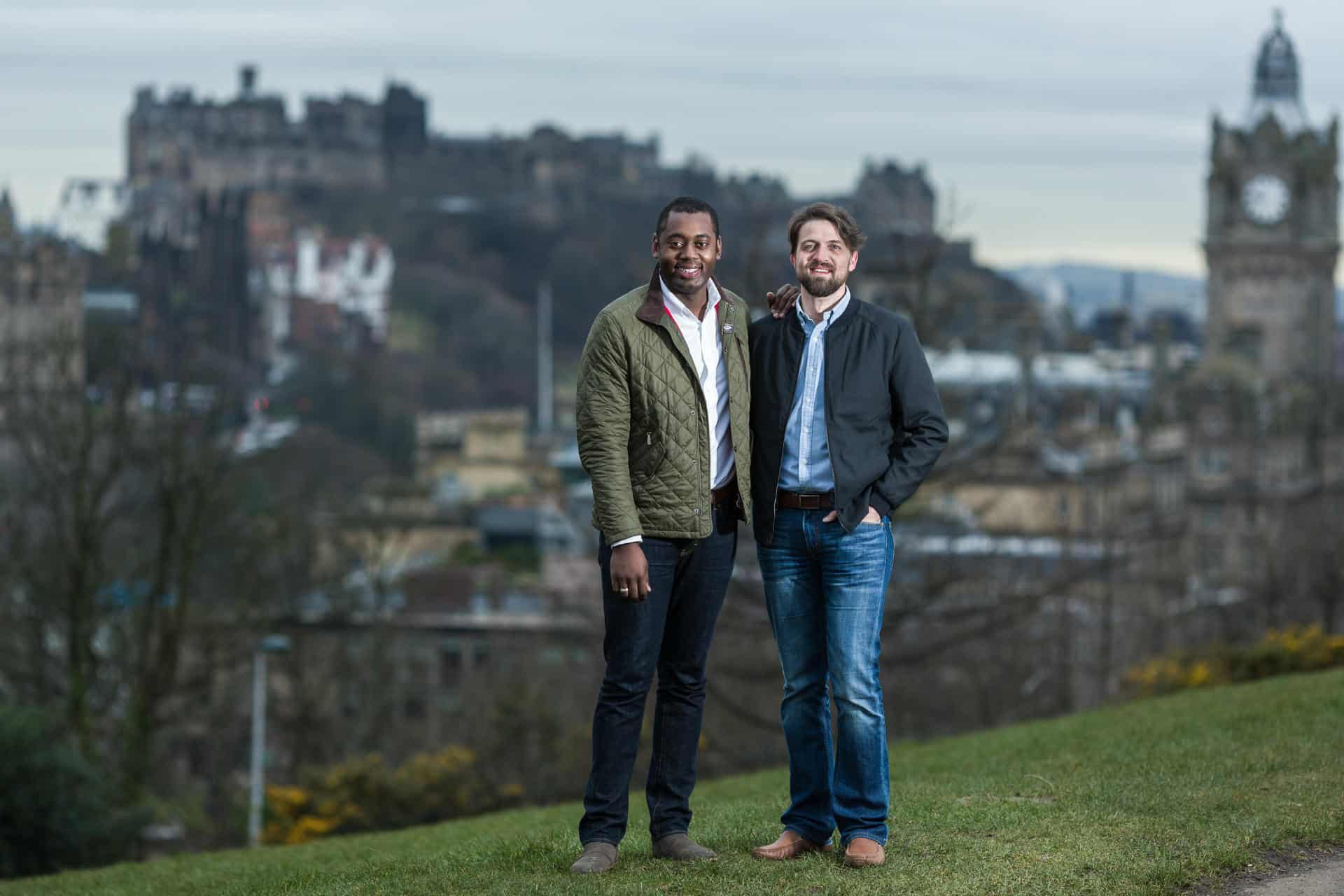 Phillip and Rick at Calton Hill: Two men stand side by side on a grassy hill, with a blurred cityscape and historic buildings in the background. One wears a green jacket, and the other wears a black jacket. They both smile at the camera.
