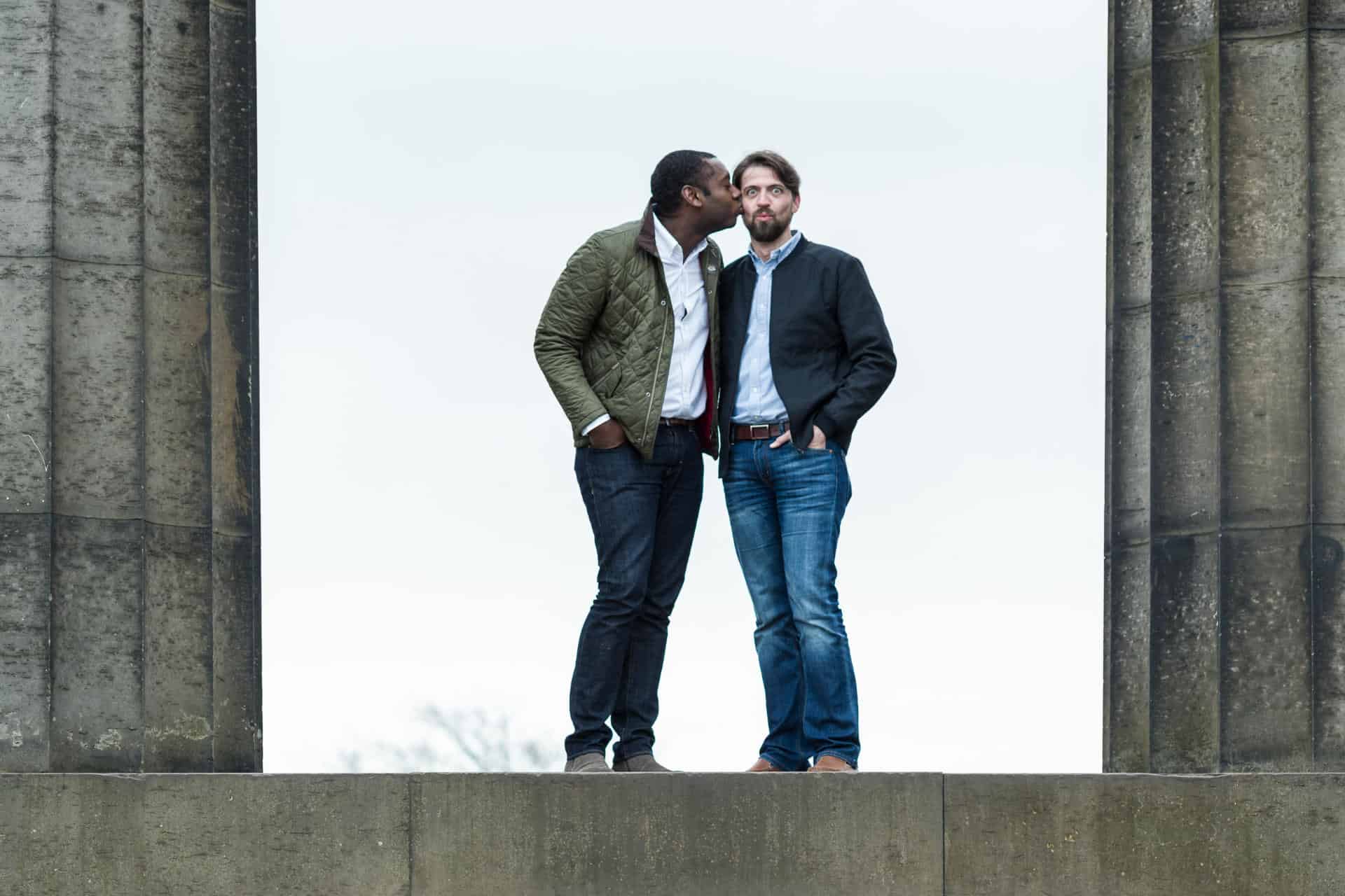 Two men stand between stone columns; one kisses the other on the temple while both face forward.