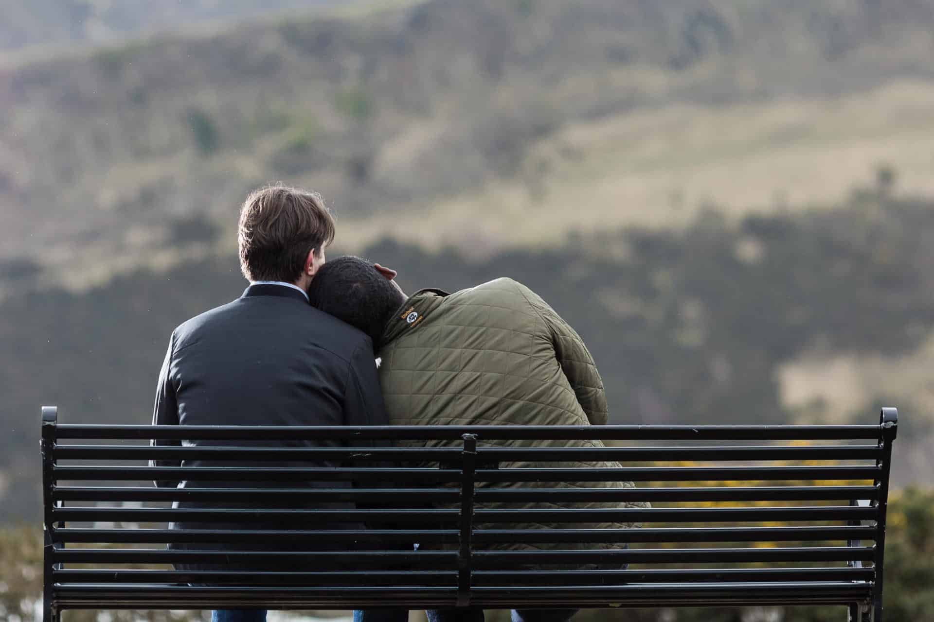 Two people sit on a black metal bench outdoors, one resting their head on the other's shoulder, facing away toward a blurred landscape of hills.