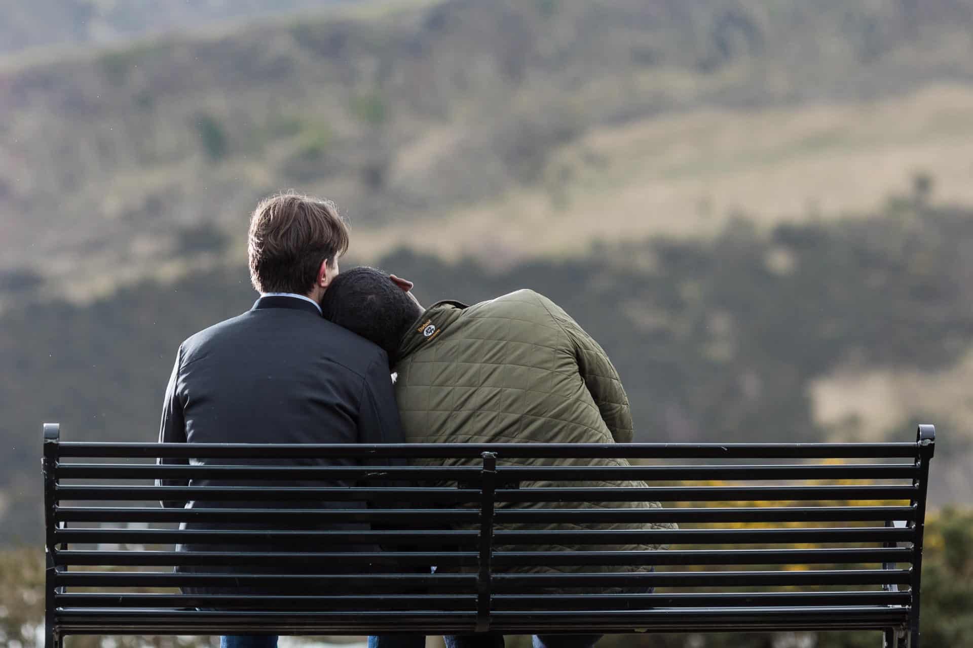 Two people sit on a black metal bench outdoors, one resting their head on the other's shoulder, facing away toward a blurred landscape of hills.