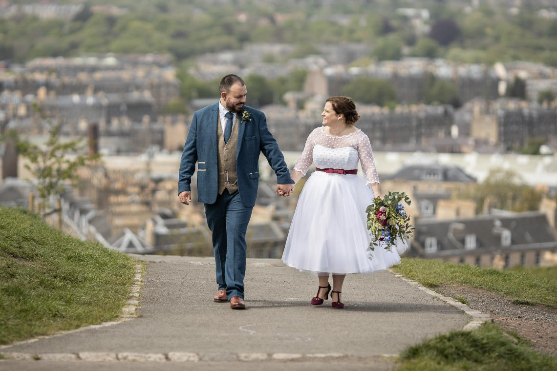 A bride and groom walk hand in hand along a path outdoors, with a bouquet in her hand and a cityscape in the background.