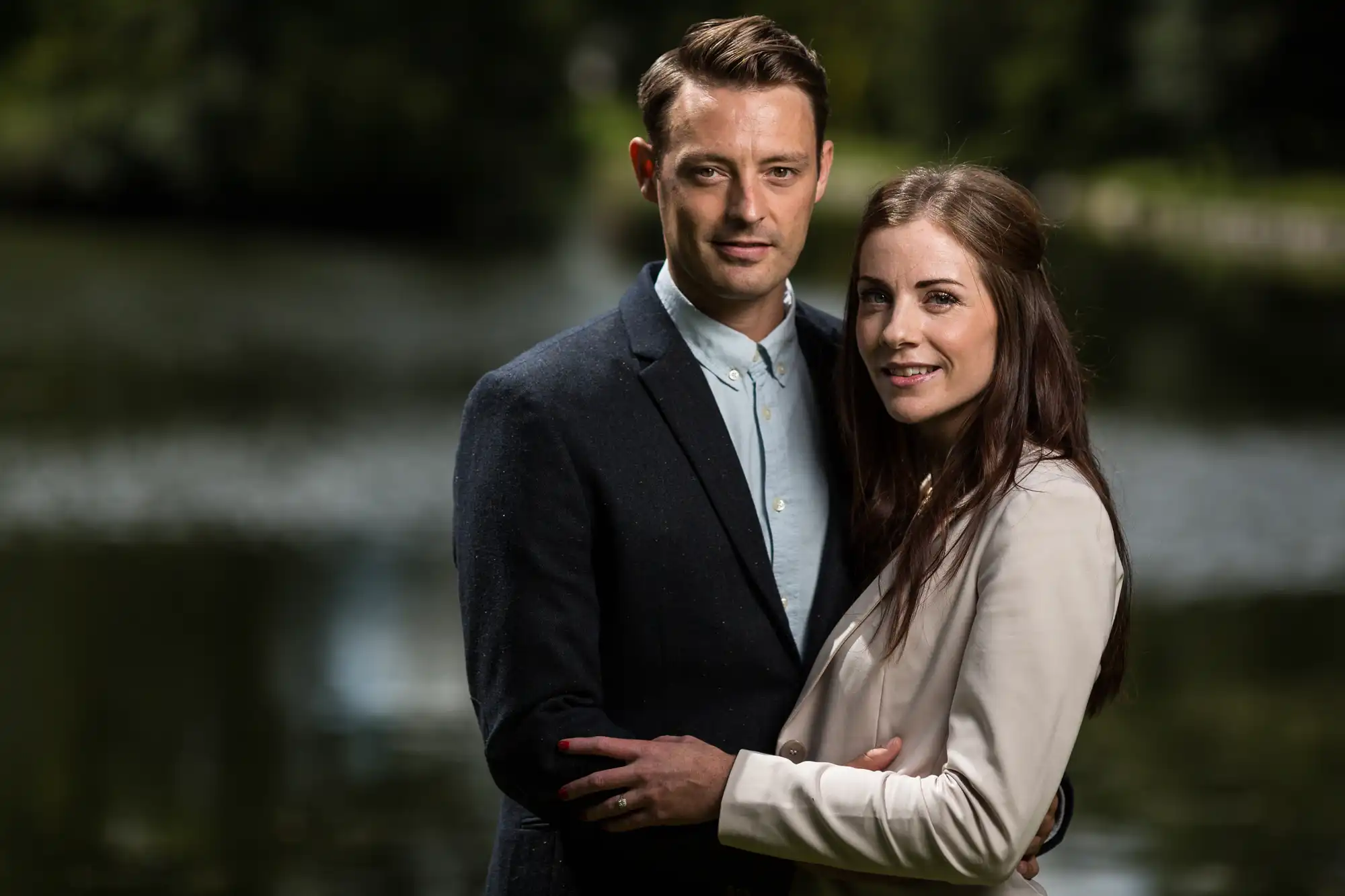 A couple embracing outdoors near a lake, the man in a blue jacket and the woman in a beige blazer, both smiling slightly at the camera.