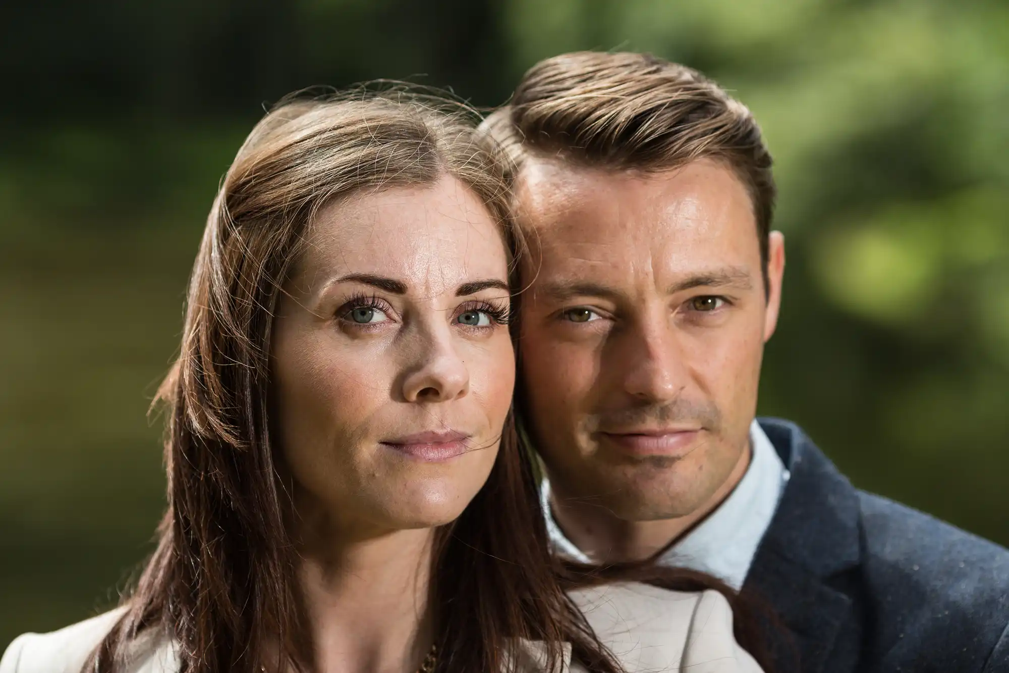 A close-up portrait of a couple, a woman and a man, outdoors with a blurred nature background, both looking seriously at the camera.