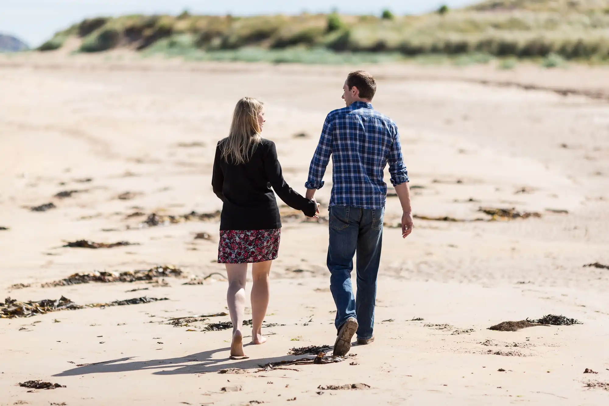 A couple holding hands while walking on a sandy beach, with the woman looking at the man, both dressed in casual attire.