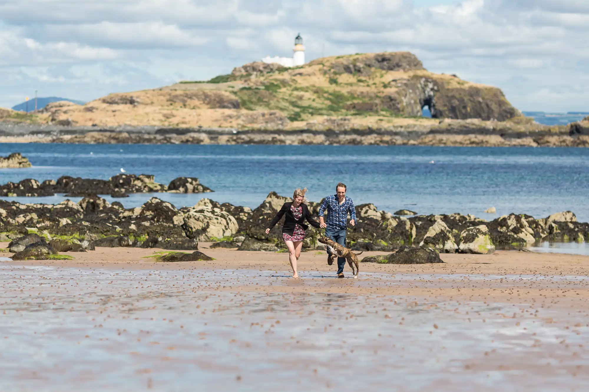 A couple and their dog running on a sandy beach, with a rocky island and lighthouse in the background.