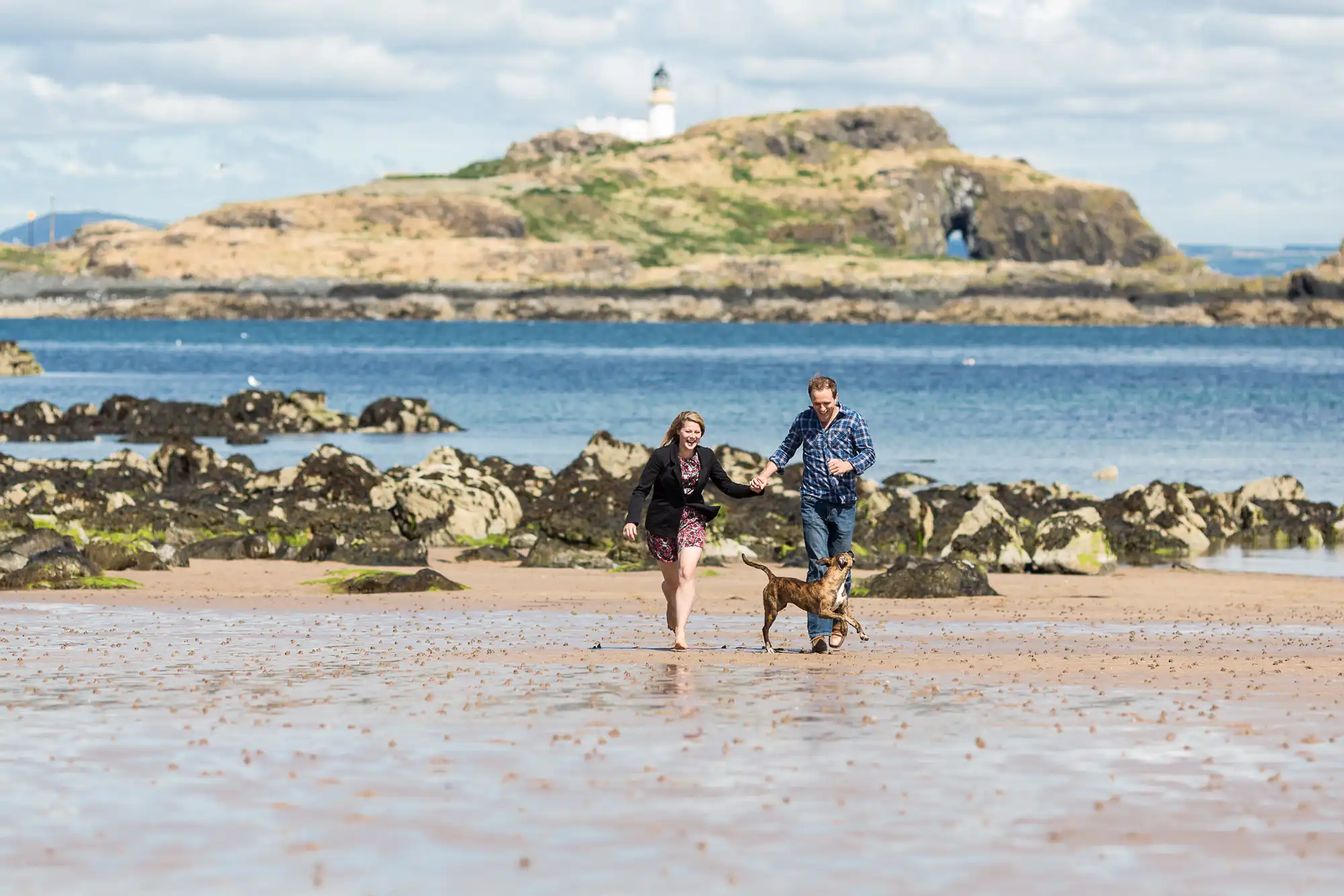 A man and a woman running on a sandy beach with a small dog, a rocky islet with a lighthouse in the background under a cloudy sky.