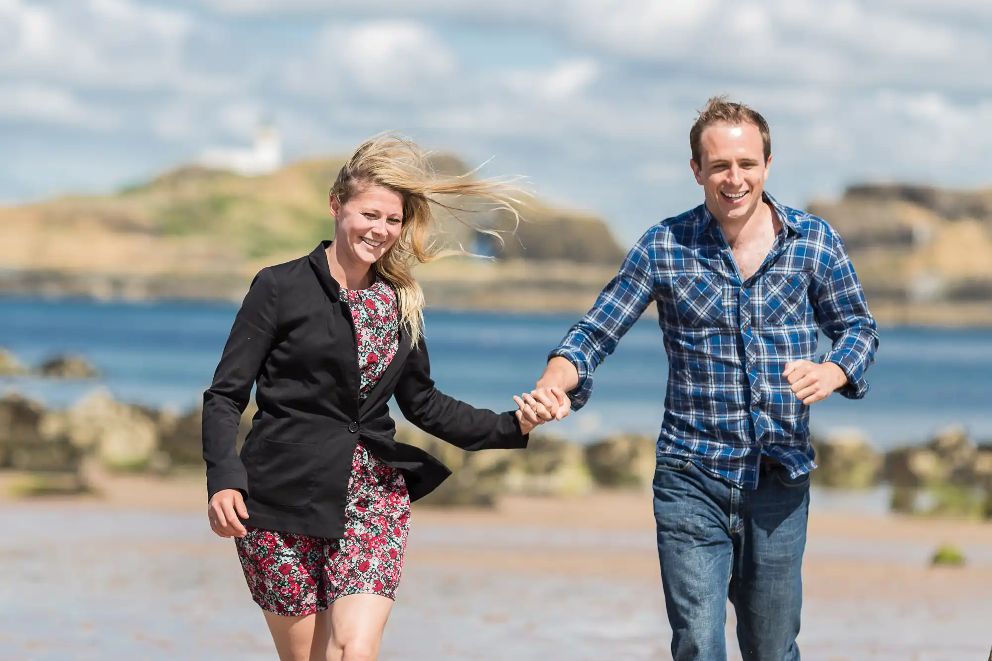 A man and a woman holding hands and smiling while running on a sandy beach with a clear blue sky and rocky island in the background.