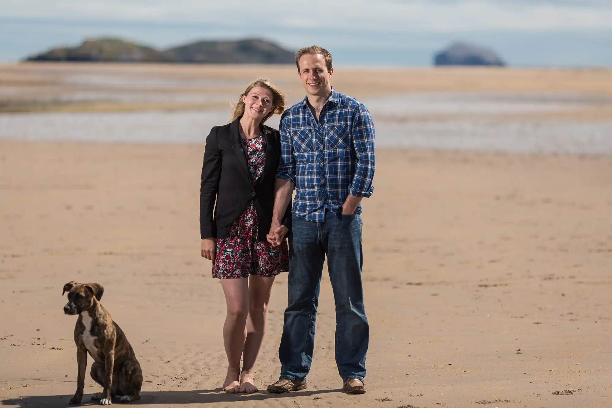 A couple standing hand-in-hand on a sandy beach, smiling at the camera, with a dog sitting beside them and islands in the background.