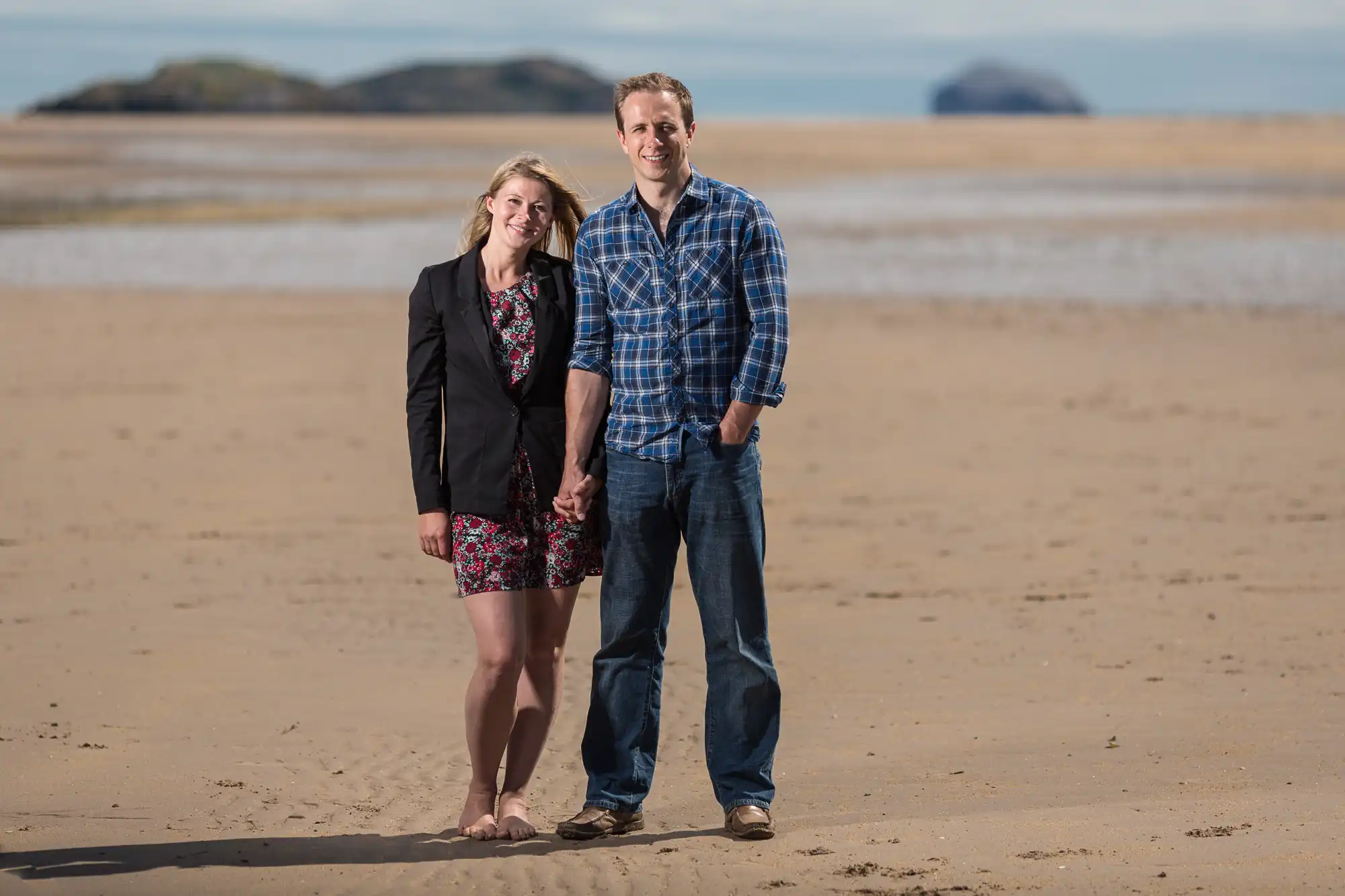 A smiling couple standing on a sandy beach with small islands visible in the distance. the woman wears a dress and blazer, and the man is in a plaid shirt and jeans.
