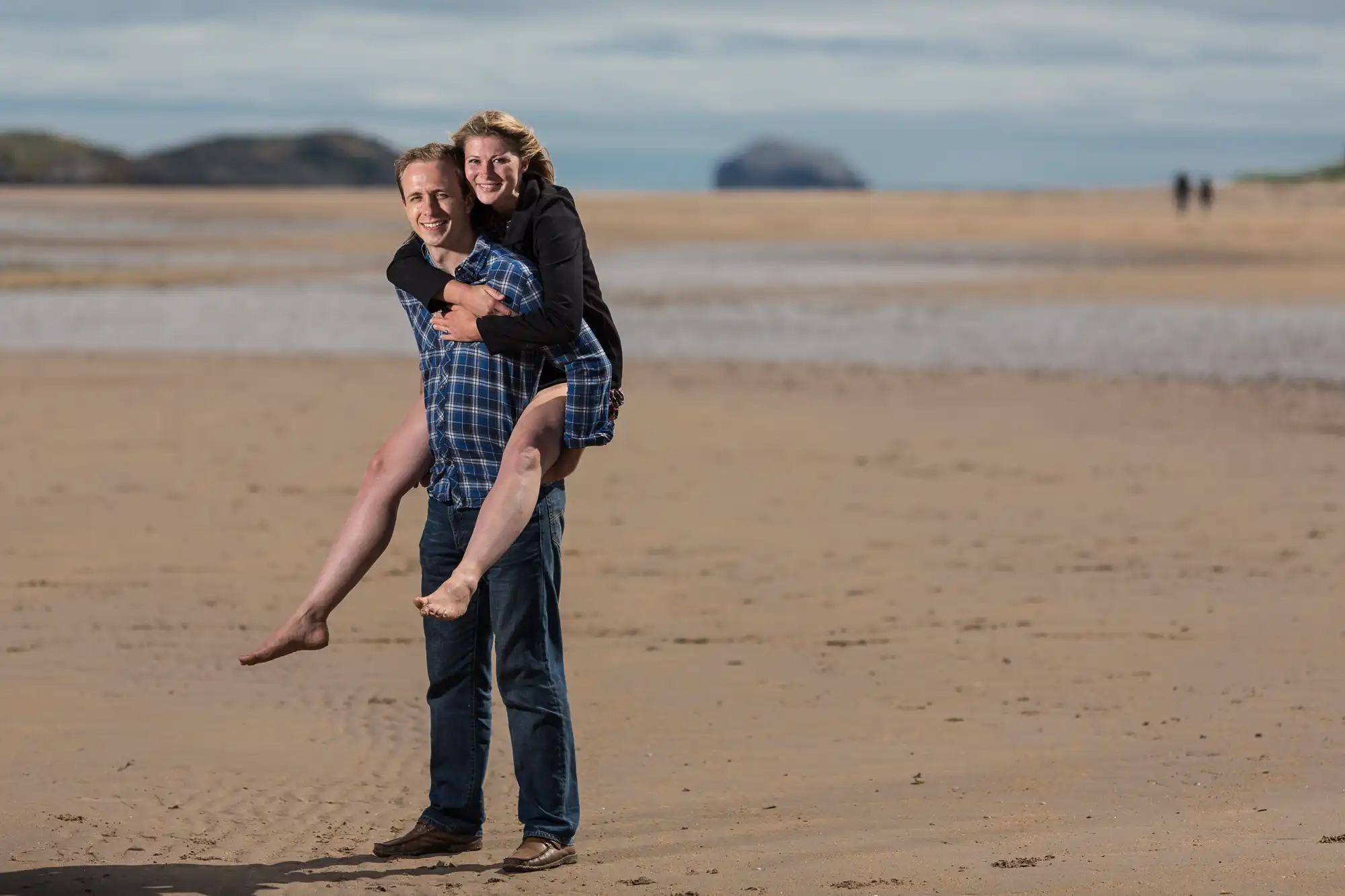 A man carries a woman piggyback on a sandy beach, both smiling, with hills in the distance under a cloudy sky.