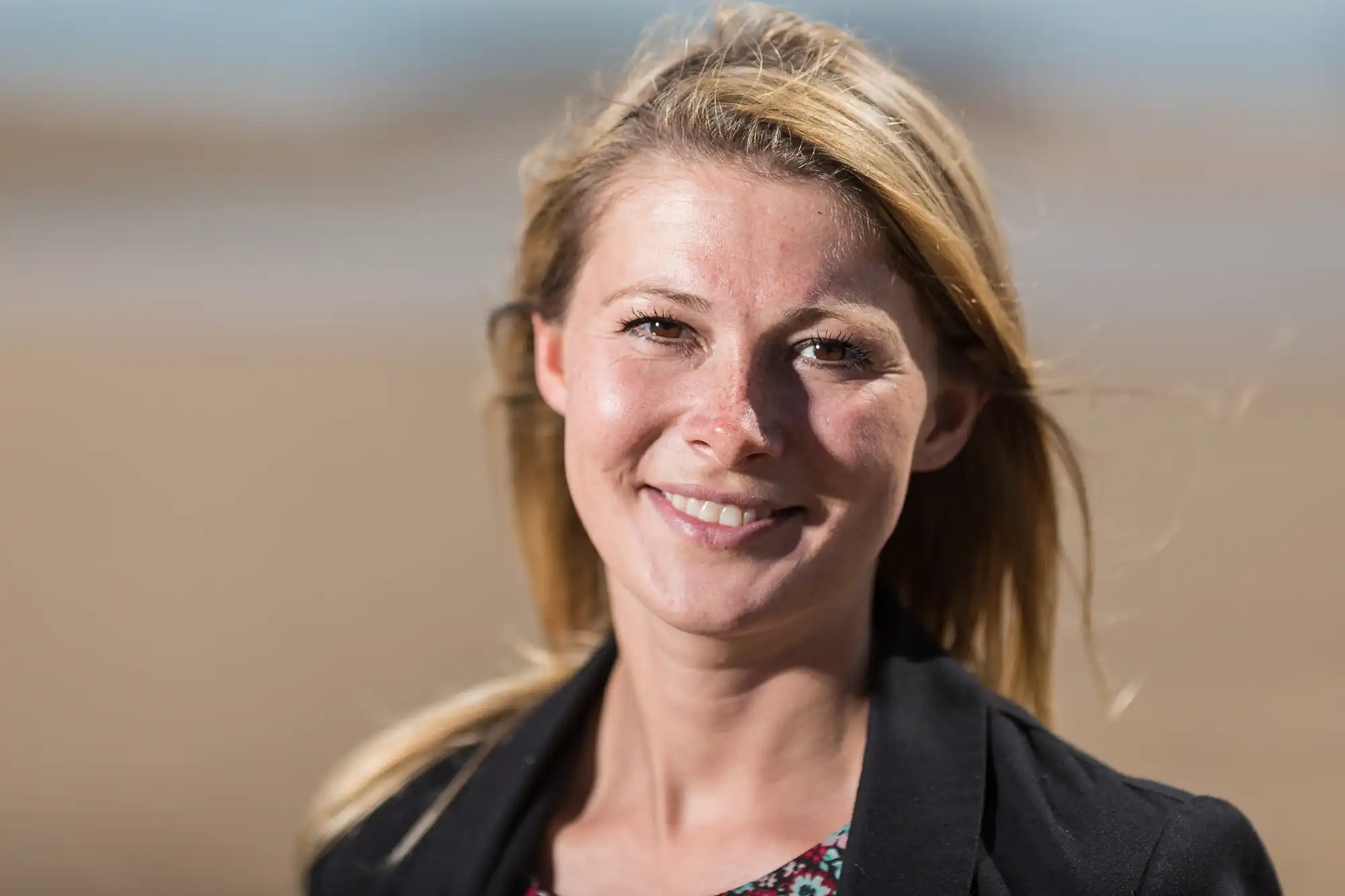 Smiling woman with blonde hair outdoors, wearing a black jacket over a floral dress, with a sandy beach blurred in the background.