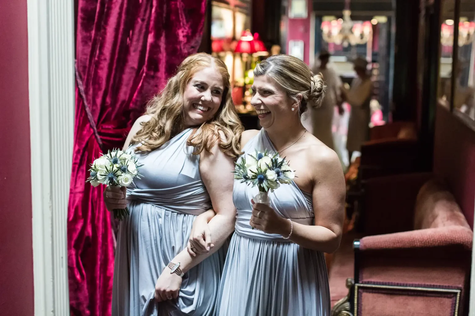 Vivid wedding photography of two bridesmaids in lavender dresses hold bouquets and smile while walking arm-in-arm down a dimly lit corridor with red curtains and chairs at Prestonfield House in Edinburgh.