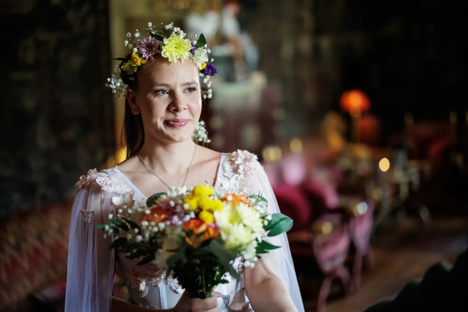 A woman wearing a flower crown and white dress holds a bouquet of flowers, standing indoors with softly lit, blurred background furniture.