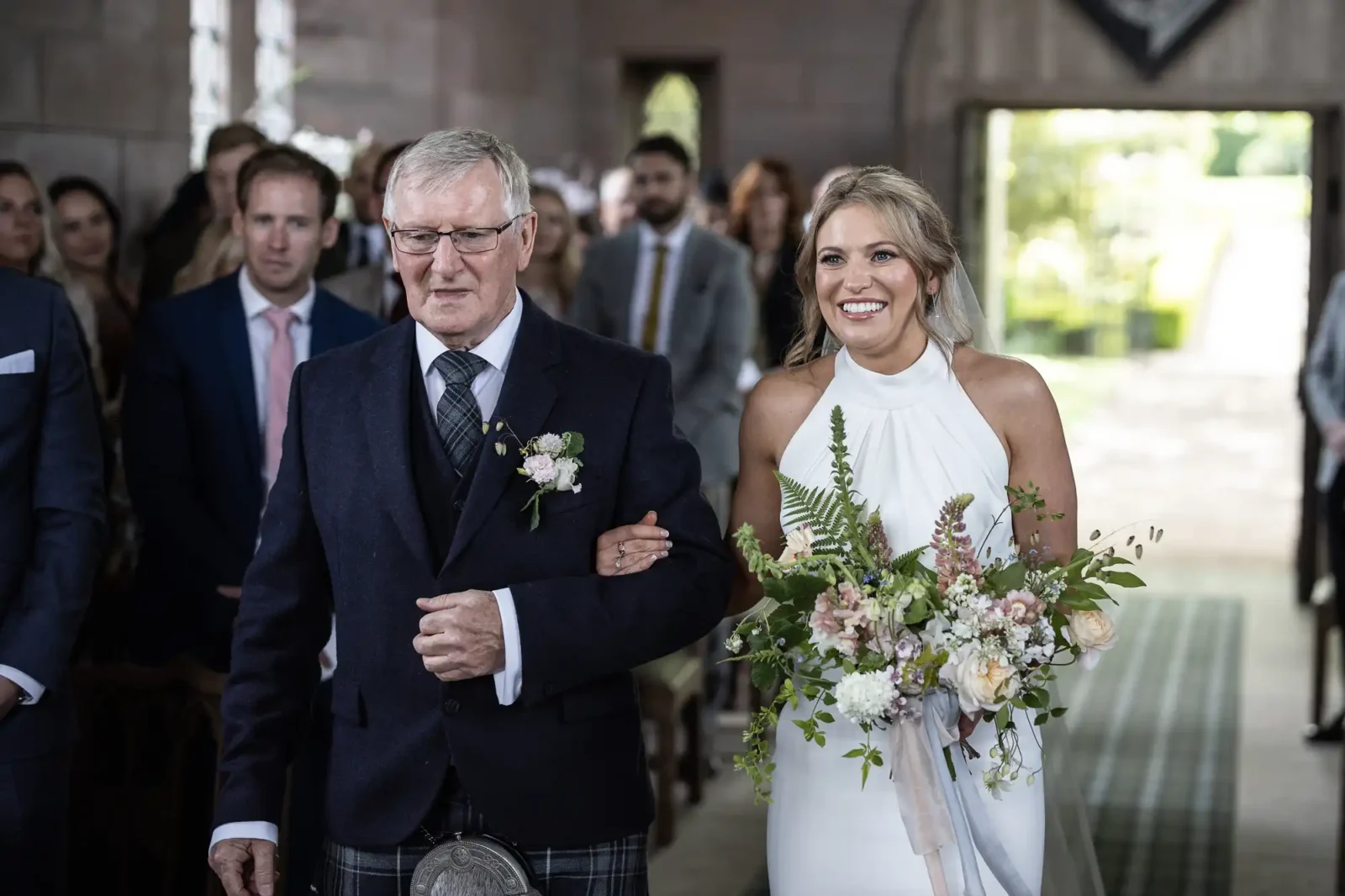 A bride in a white halter neck dress smiling as she walks down the aisle with an older man in a suit, both holding a bouquet, in a room with guests watching.