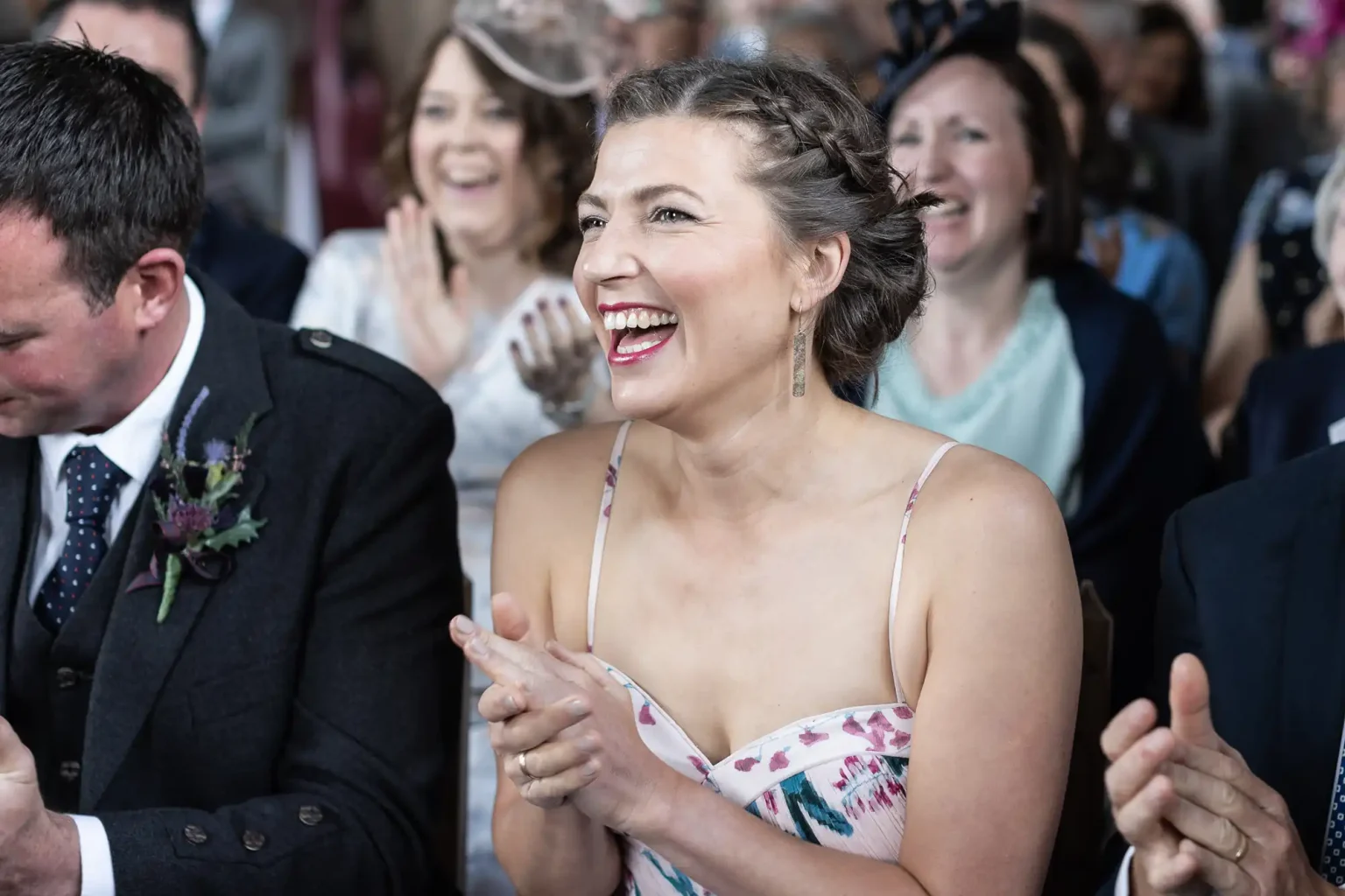 A joyful woman in a floral dress clapping at a wedding ceremony by the photographer, surrounded by other guests.
