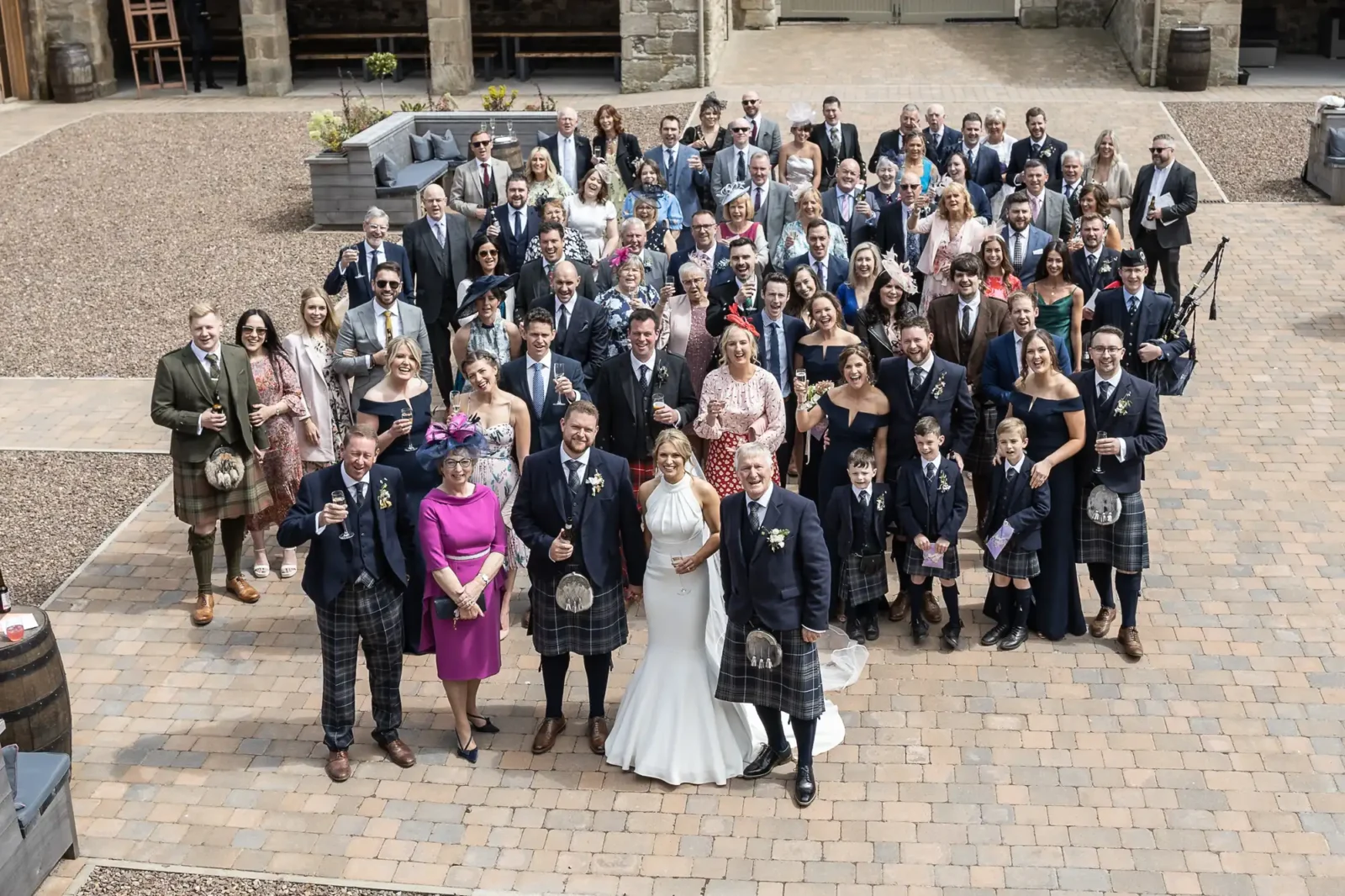 A large wedding party posing with the bride and groom, who are in the center, in a courtyard setting. Most men wear kilts and women are in dresses.