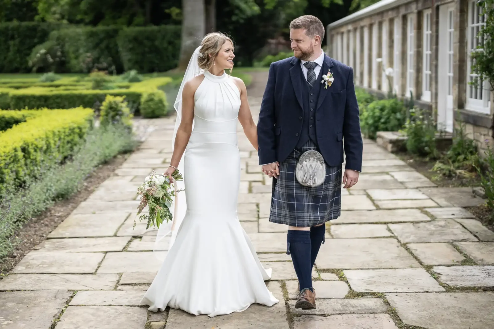 Rosebery Steading wedding photographer portrait of Gillian and Nick walking together in the grounds; A bride in a white dress and a groom in a kilt holding hands and walking along a garden path, smiling at each other.