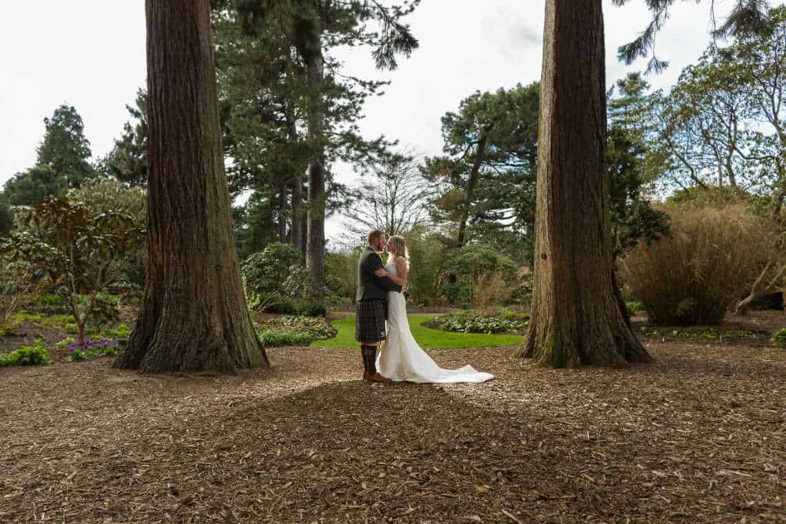 A couple wearing wedding attire stands in an embrace between two large trees in a forested area - Royal Botanic Garden Edinburgh - Fiona and Chris