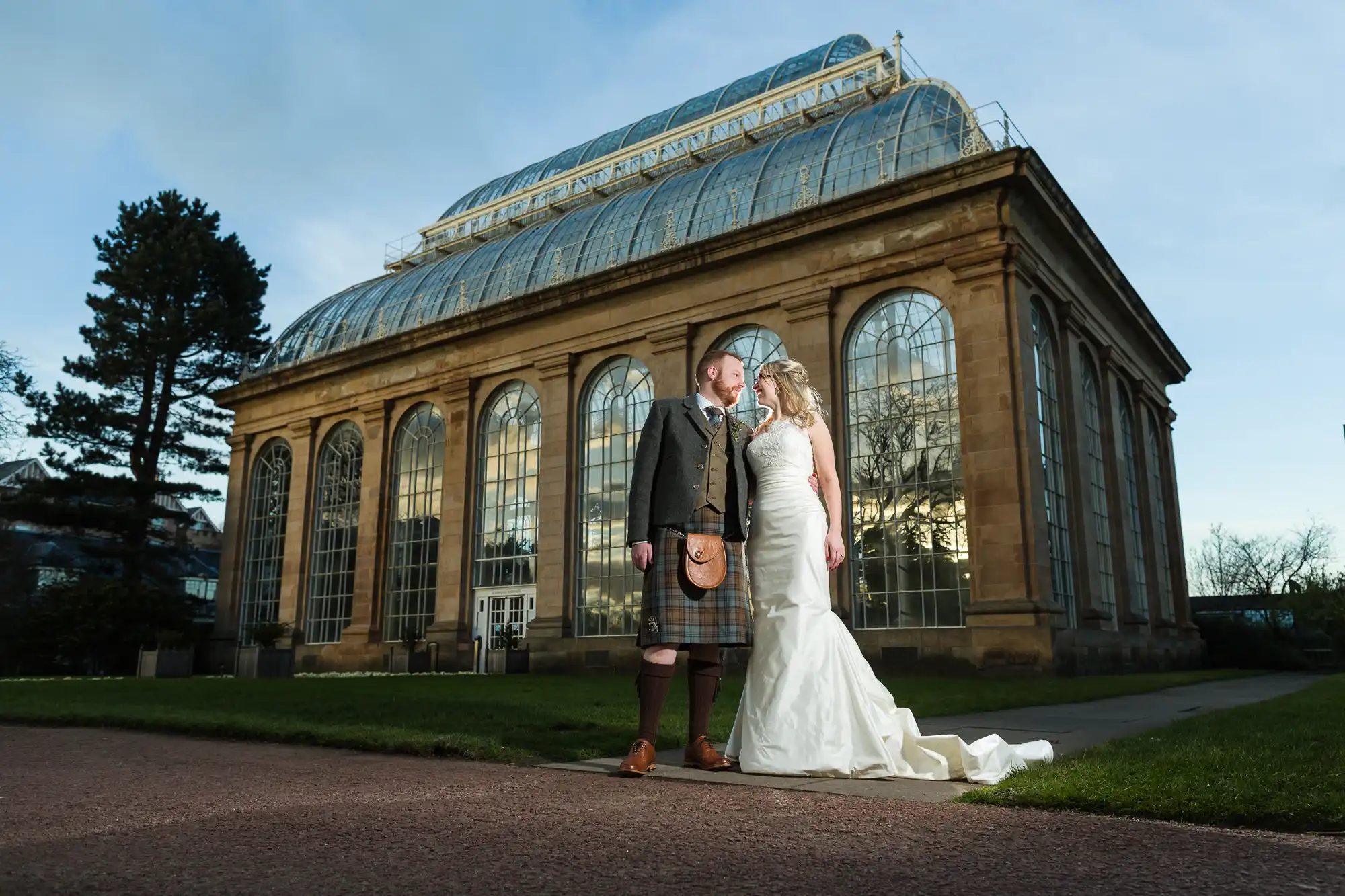Royal Botanic Garden Edinburgh wedding photography - natural floral backdrop photos. A couple in wedding attire stands in front of a large, glass-domed building. The groom is in a kilt and jacket, while the bride wears a strapless white gown with a long train.
