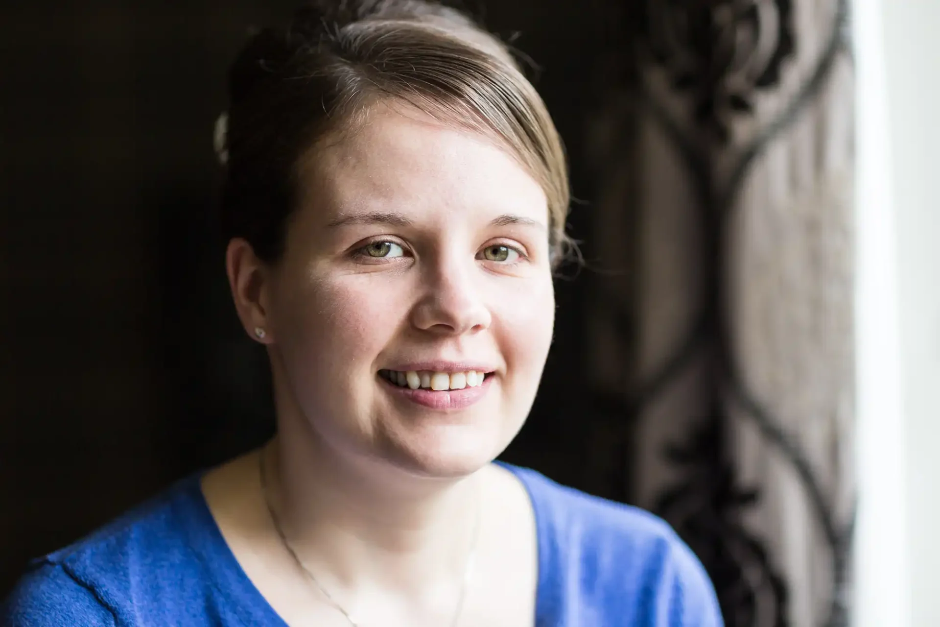 A woman with short brown hair and a blue top smiling softly by a window with natural light illuminating her face.