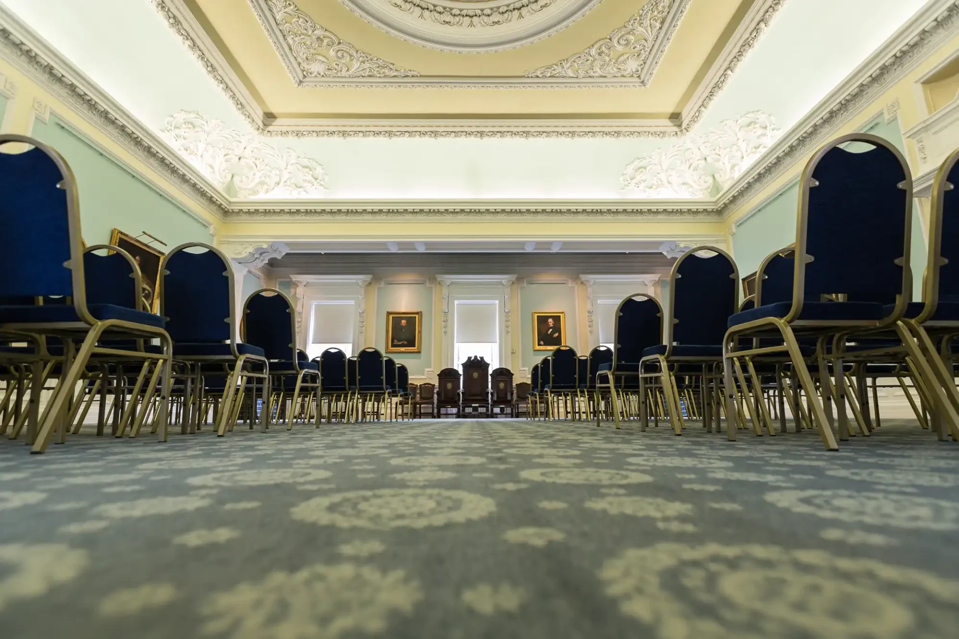 Elegant room with ornate ceiling, blue chairs arranged in rows, and framed paintings on the walls.
