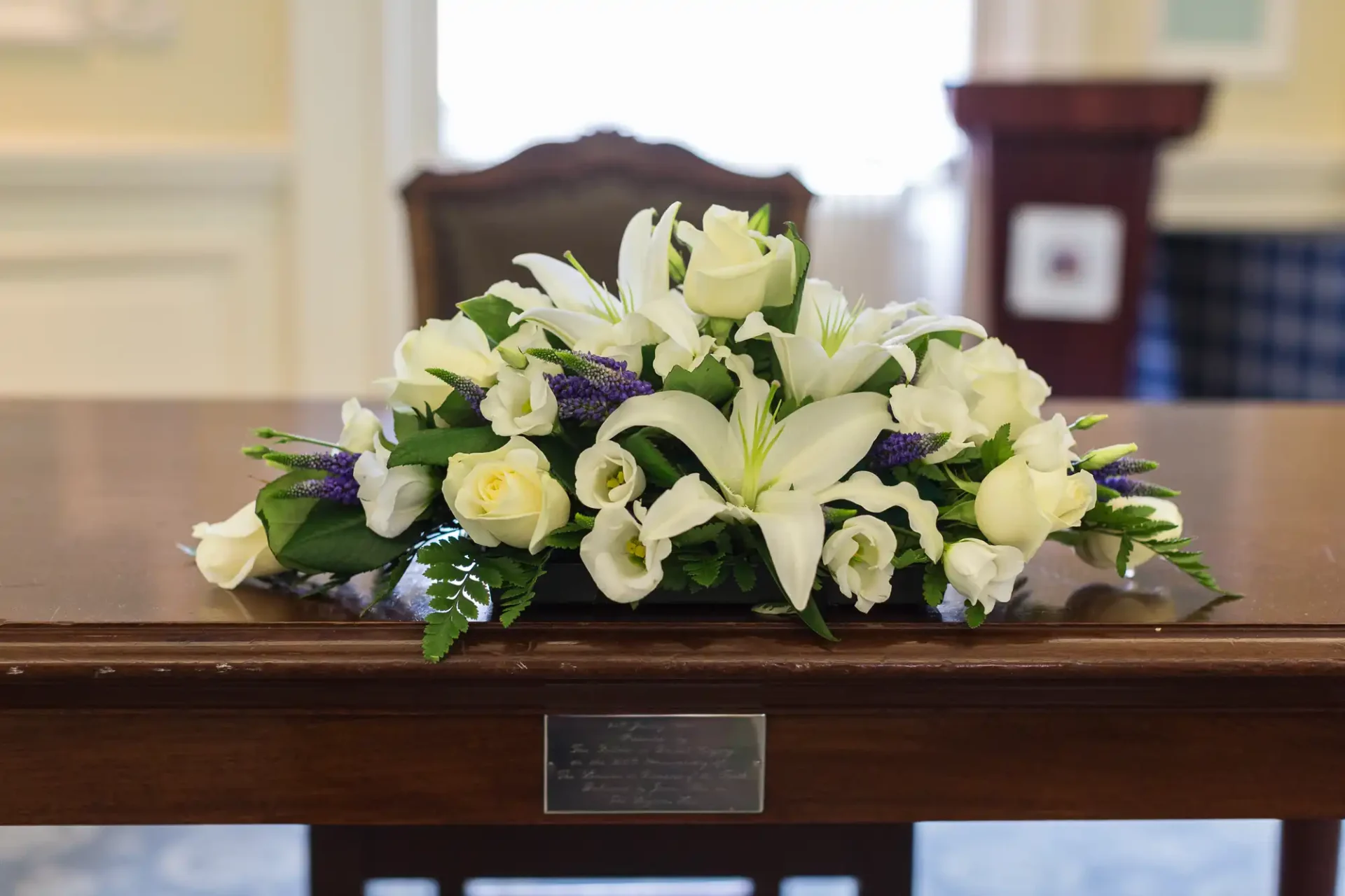 A floral arrangement with white roses and lilies on a polished wooden table in a room with cream walls and a dark chair in the background.