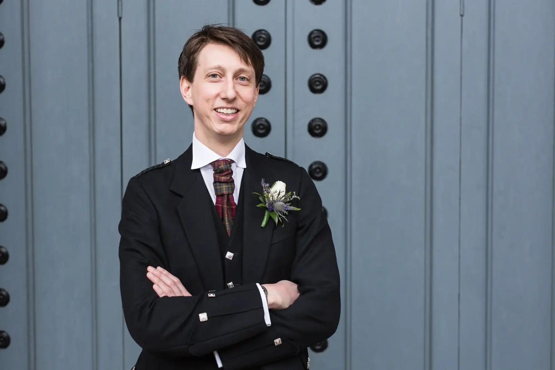 A man in a formal black suit with a white boutonniere smiles while standing with his arms crossed in front of a blue door.