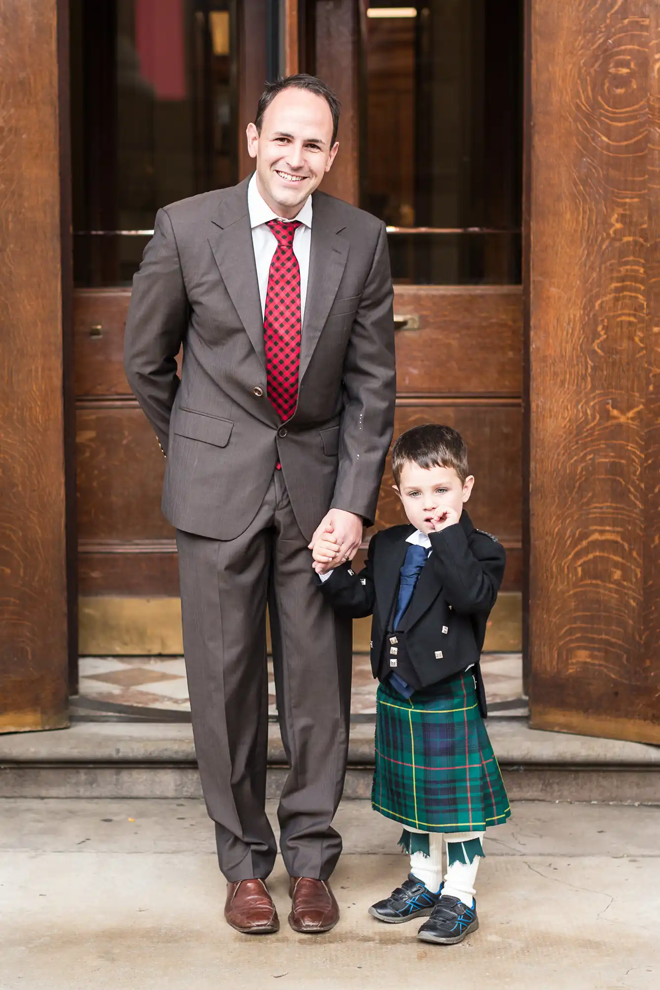 A man in a gray suit and a young boy in a kilt and jacket holding hands in front of a wooden door.