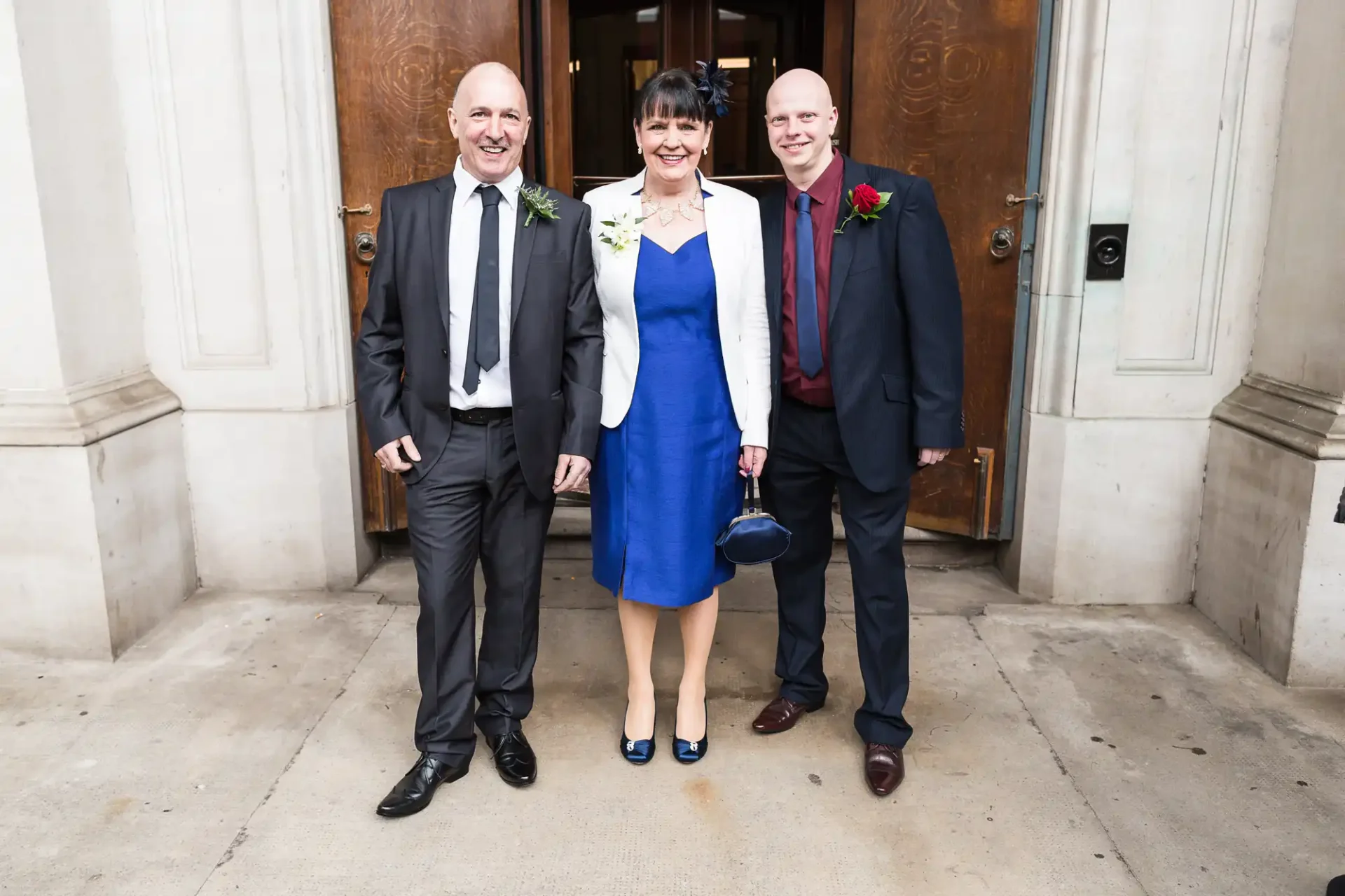 Three people dressed in formal attire with boutonnieres posing for a photo outside an elegant building entrance.