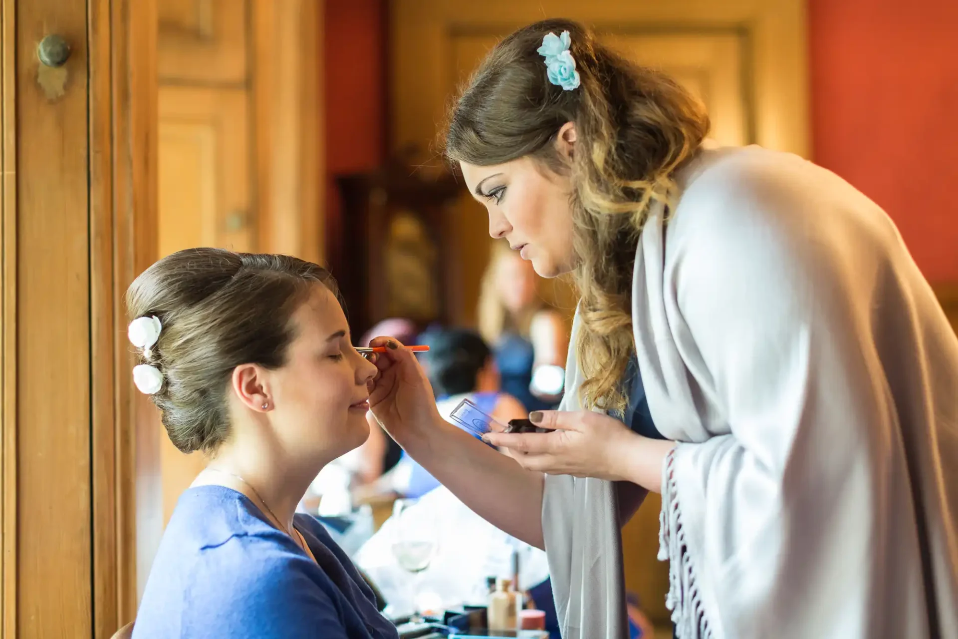 A woman applying makeup to another woman in a warmly lit room.