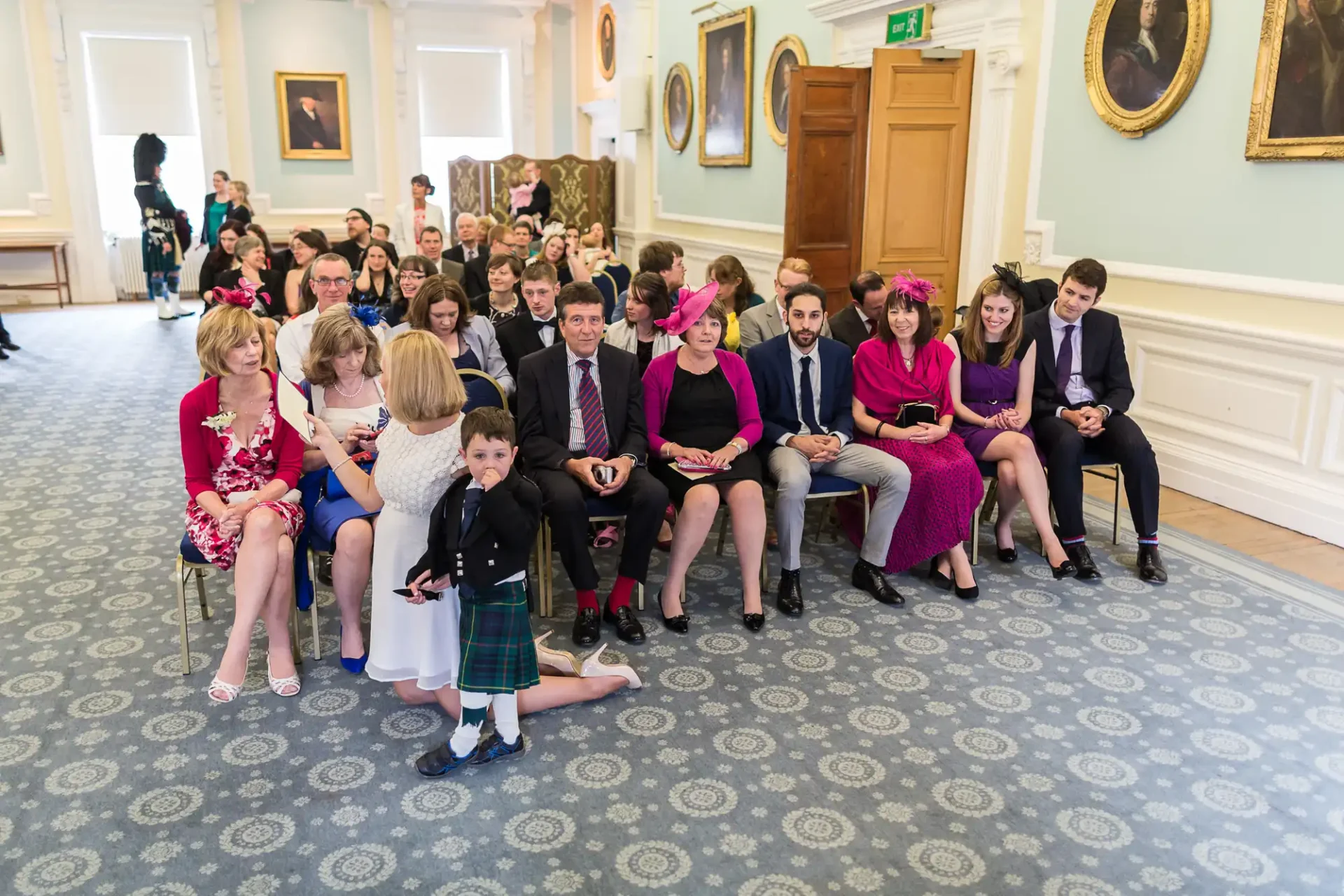 Guests in formal attire seated in rows at a wedding ceremony indoors, with portraits on the wall and a boy in a kilt in the foreground.