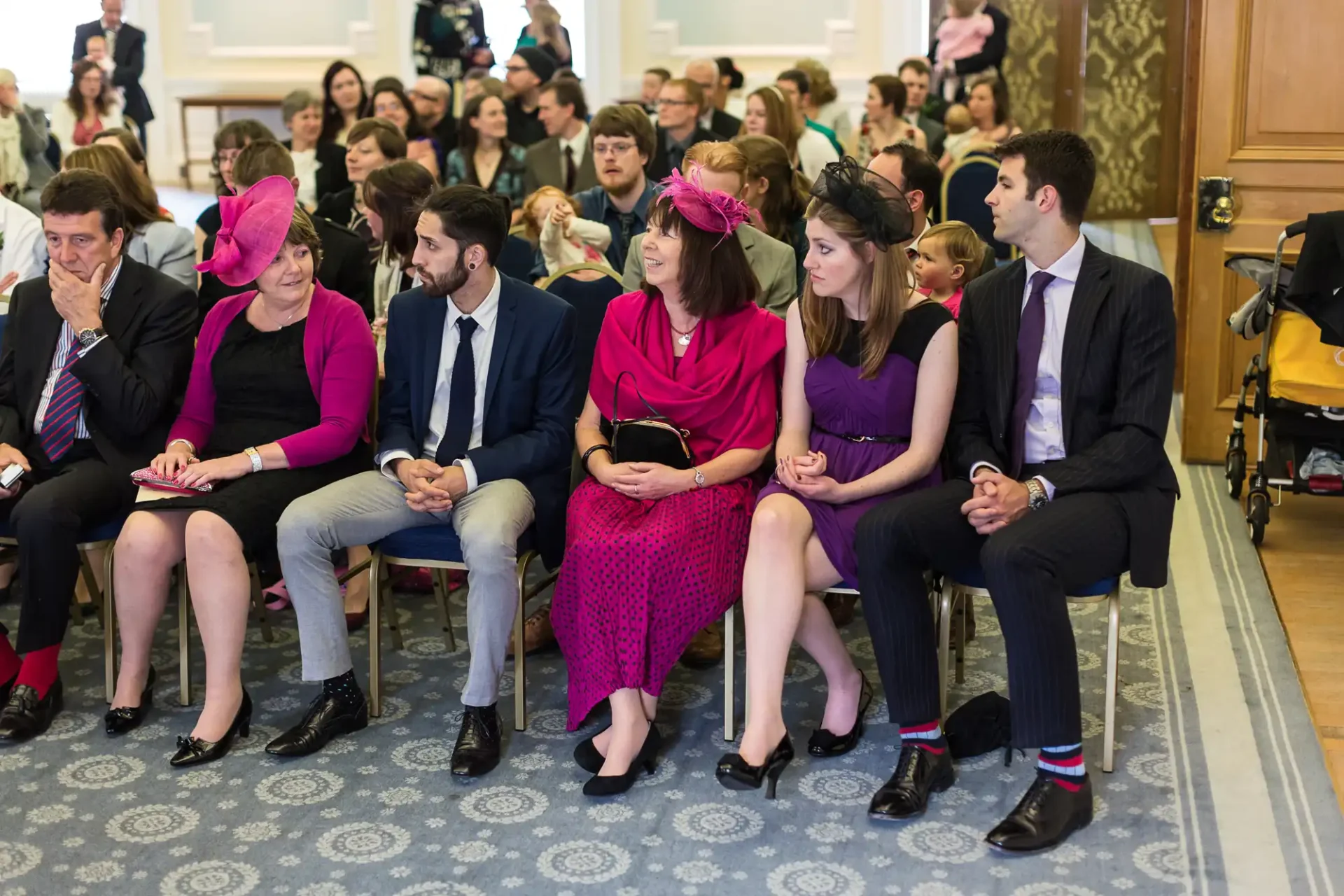 Group of adults in formal attire seated at a ceremony, with some women wearing colorful hats.