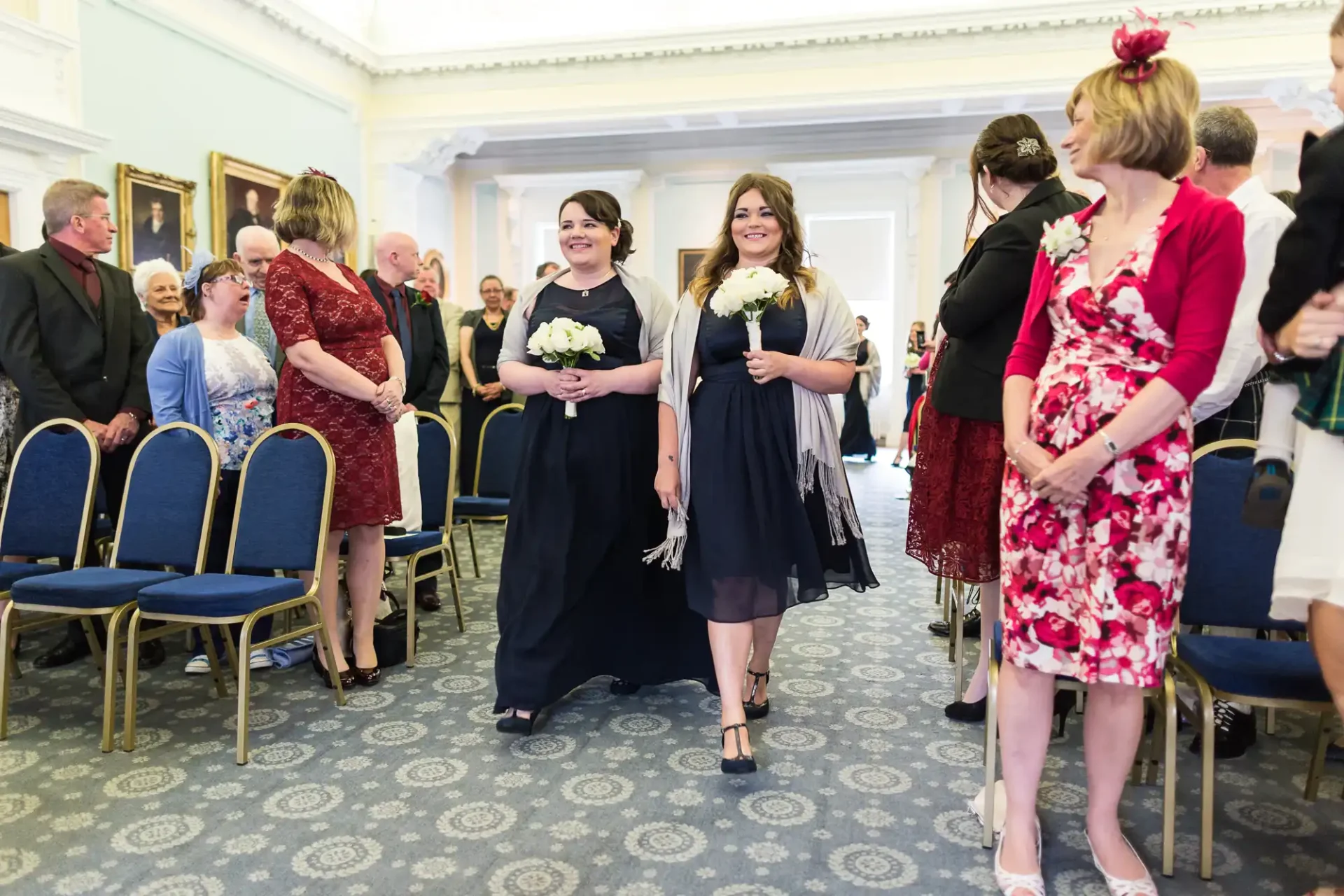 Three bridesmaids holding bouquets walk down the aisle in front of seated guests in an elegantly decorated room.