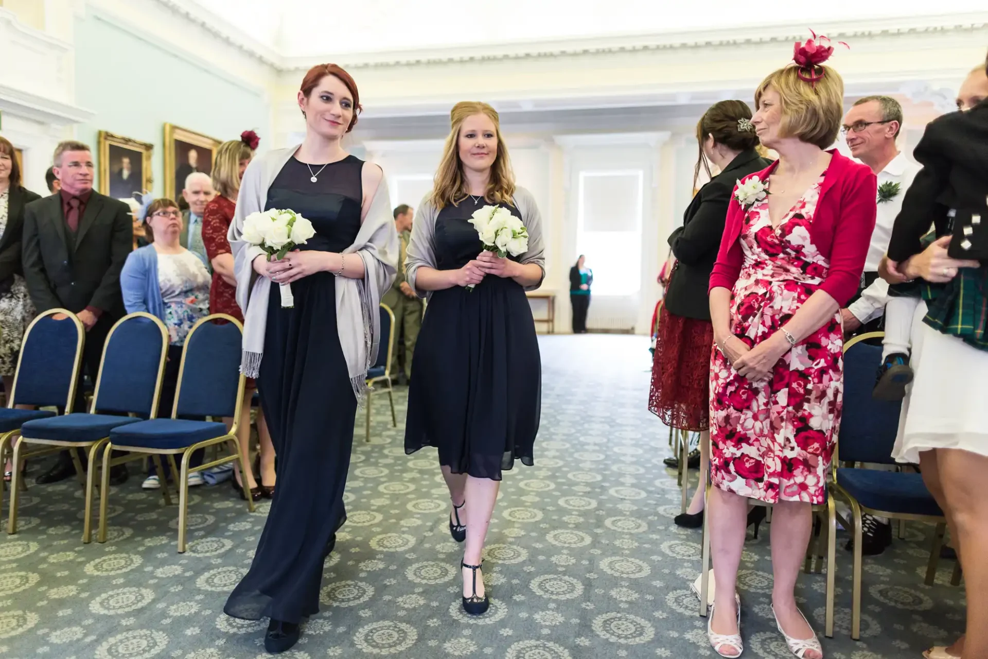 Two bridesmaids holding bouquets walk down the aisle in a decorated room with guests standing and watching.