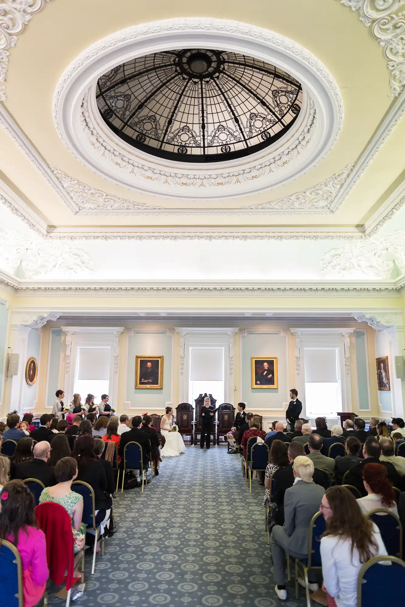 Interior of an elegant hall during a wedding ceremony, featuring ornate ceiling details, portraits on the walls, and guests seated facing a couple at the front.