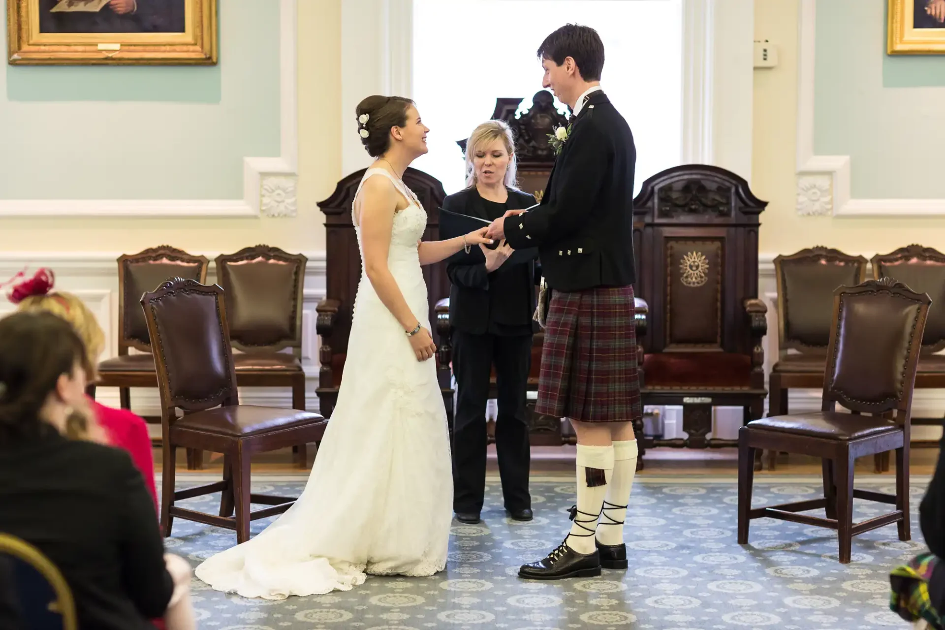 A bride and groom in traditional attire exchanging rings during a wedding ceremony indoors while the officiant and guests watch.