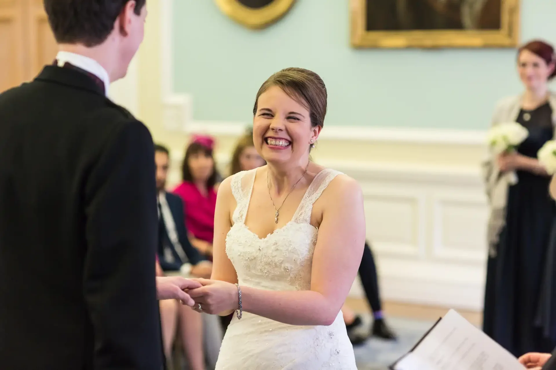 A bride in a white dress smiling joyfully at the groom during a wedding ceremony, with guests in the background.