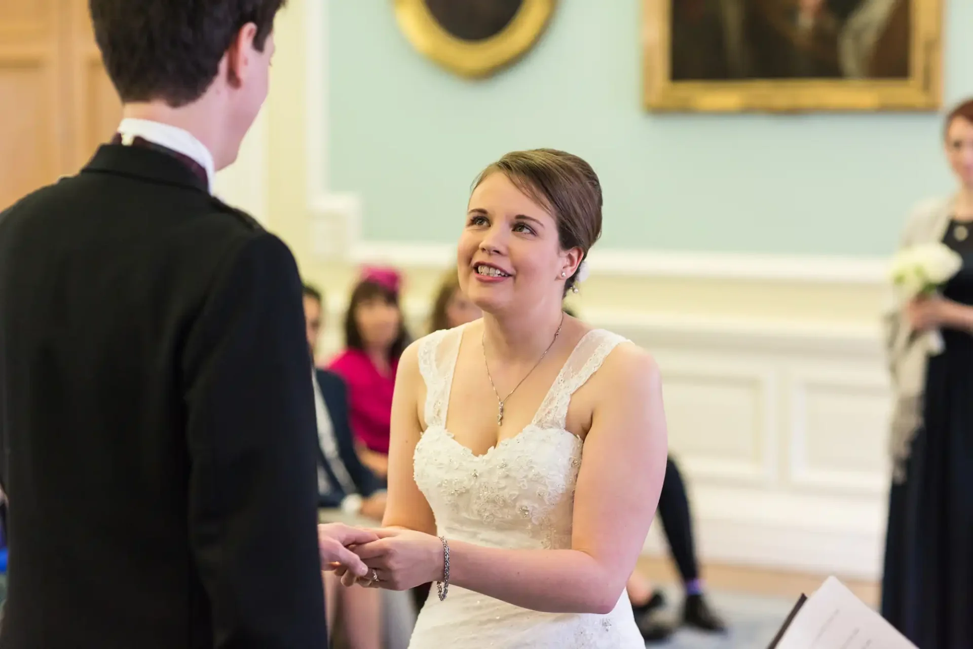 Bride in a white dress exchanging vows with groom, both standing in a room with portraits on the walls, viewed from the side.