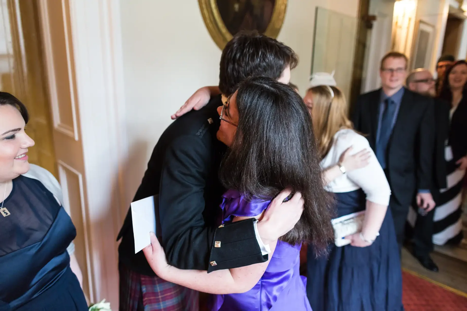 A woman in a purple dress embracing a man in a suit at a formal event, with observers in the background.