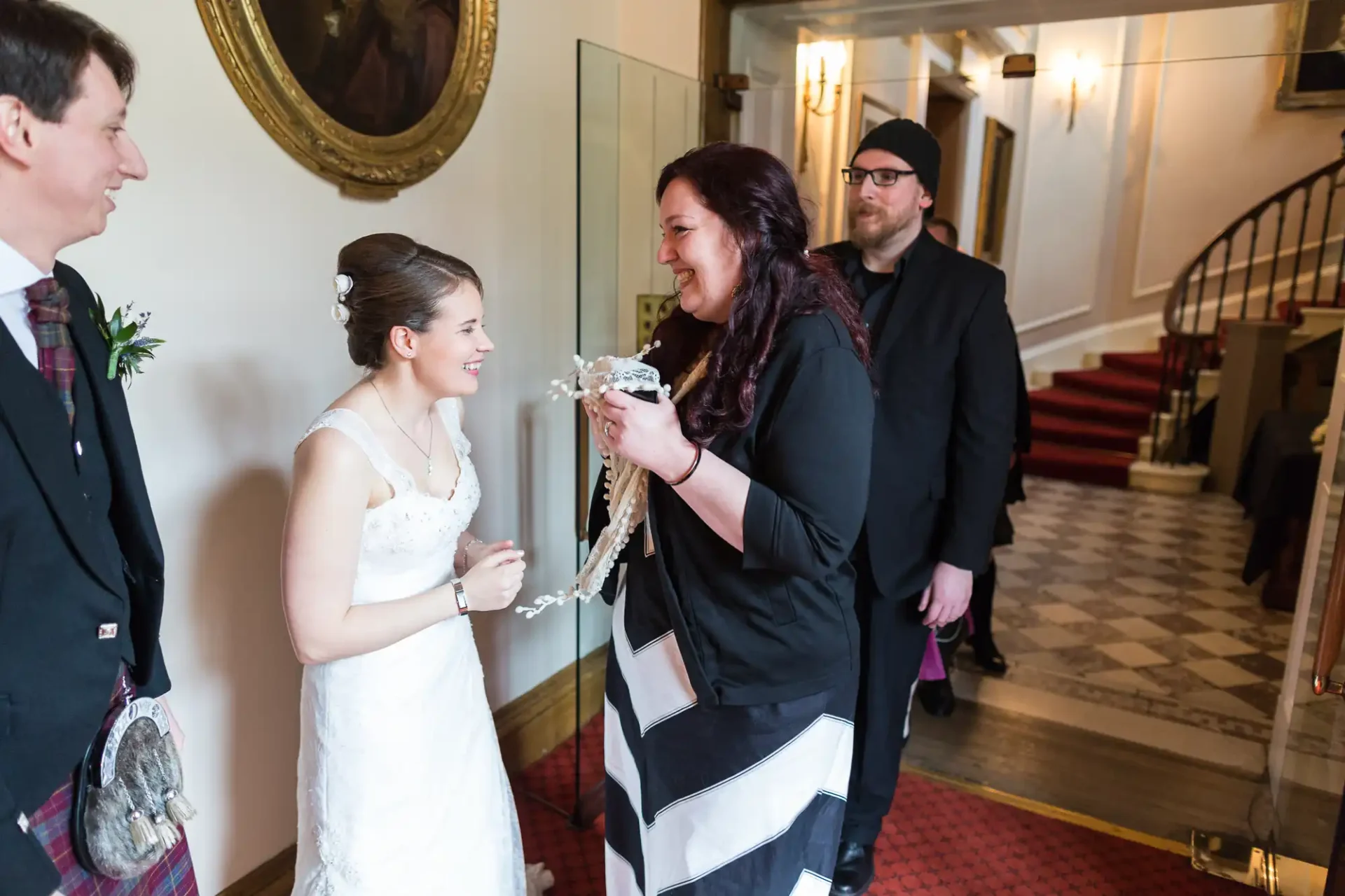 A bride in a white dress smiles while receiving a decorative item from a woman in a black and white dress in a hotel lobby, as two men observe.