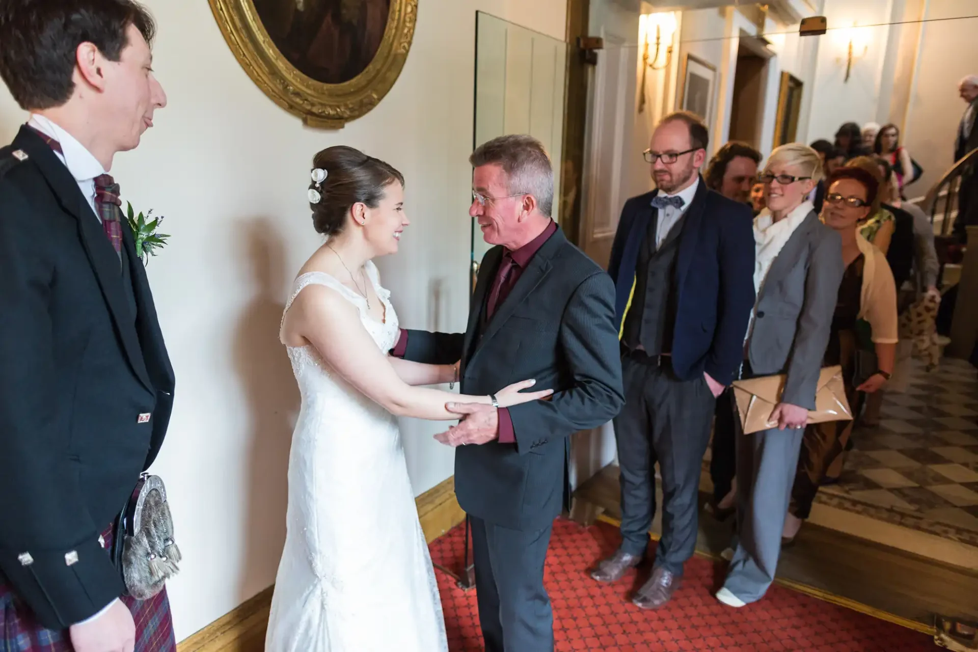 A bride in a white dress converses with a man in a dark suit in a hallway, as guests in varied attire observe nearby.