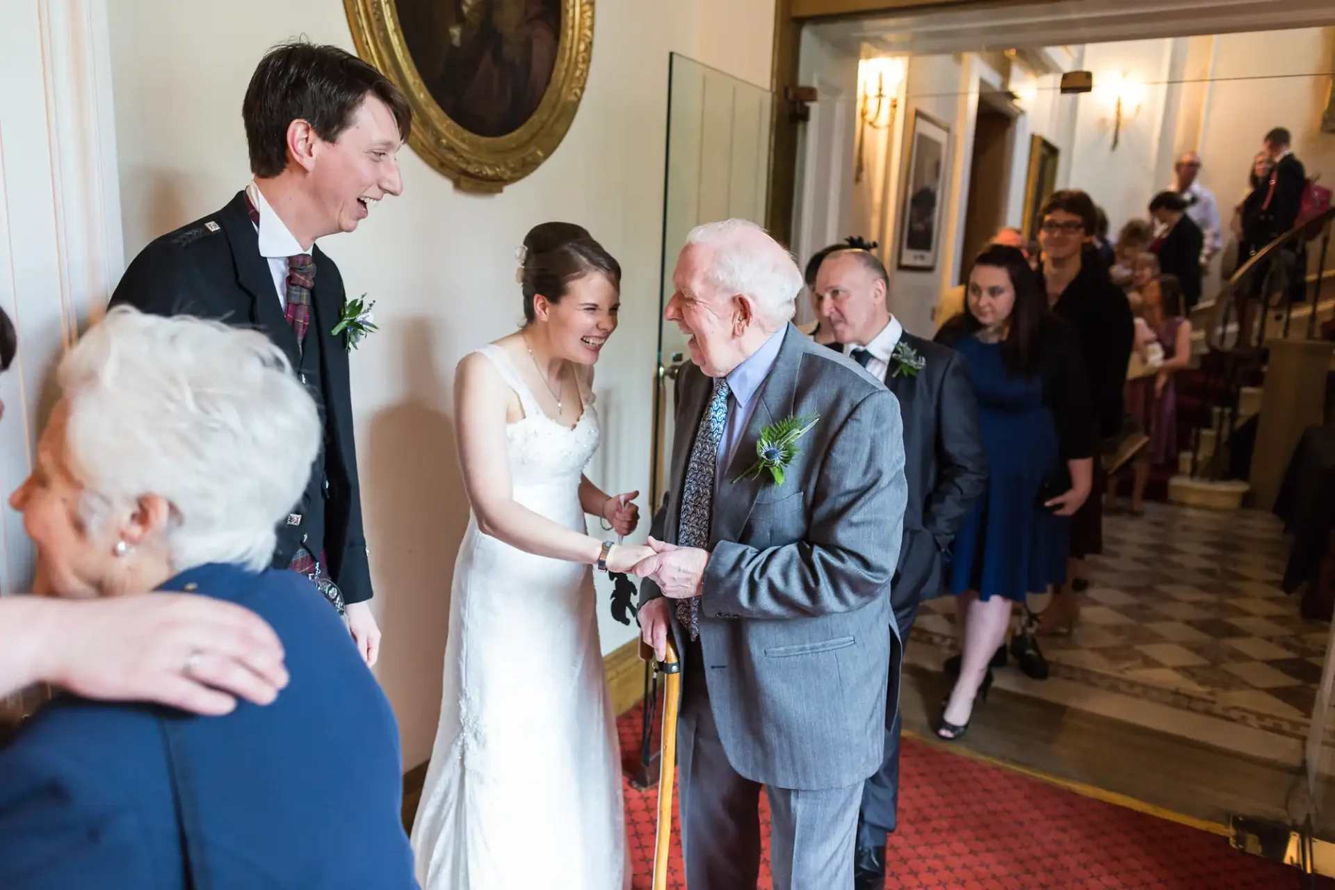 A bride and groom greet an elderly man at a wedding reception, surrounded by guests in a warmly lit hall.