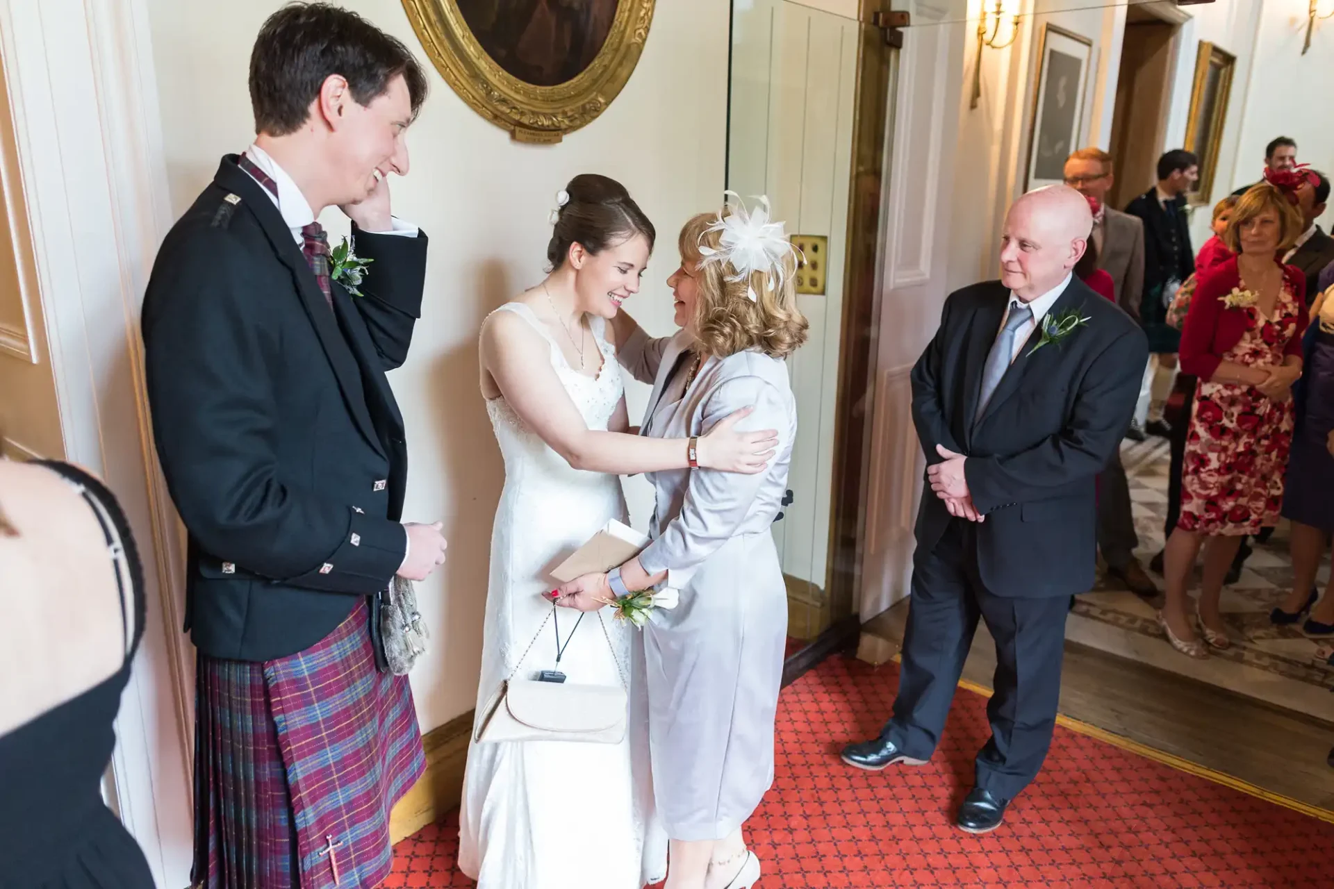 A bride and groom in formal wedding attire greet guests in a warmly lit room, showing smiles and engaged in a conversation.