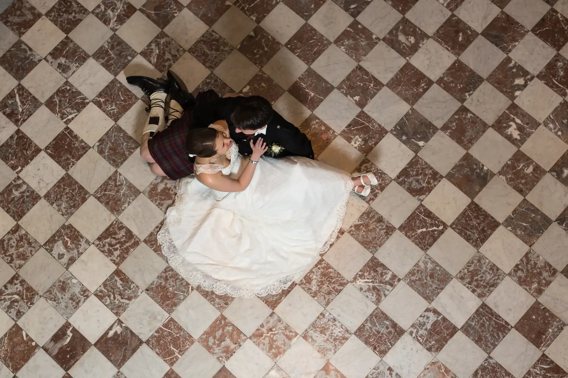 A bride in a white dress and a groom in a black suit lie on a checkered floor, sharing a kiss, with the bride's dress spread around them.
