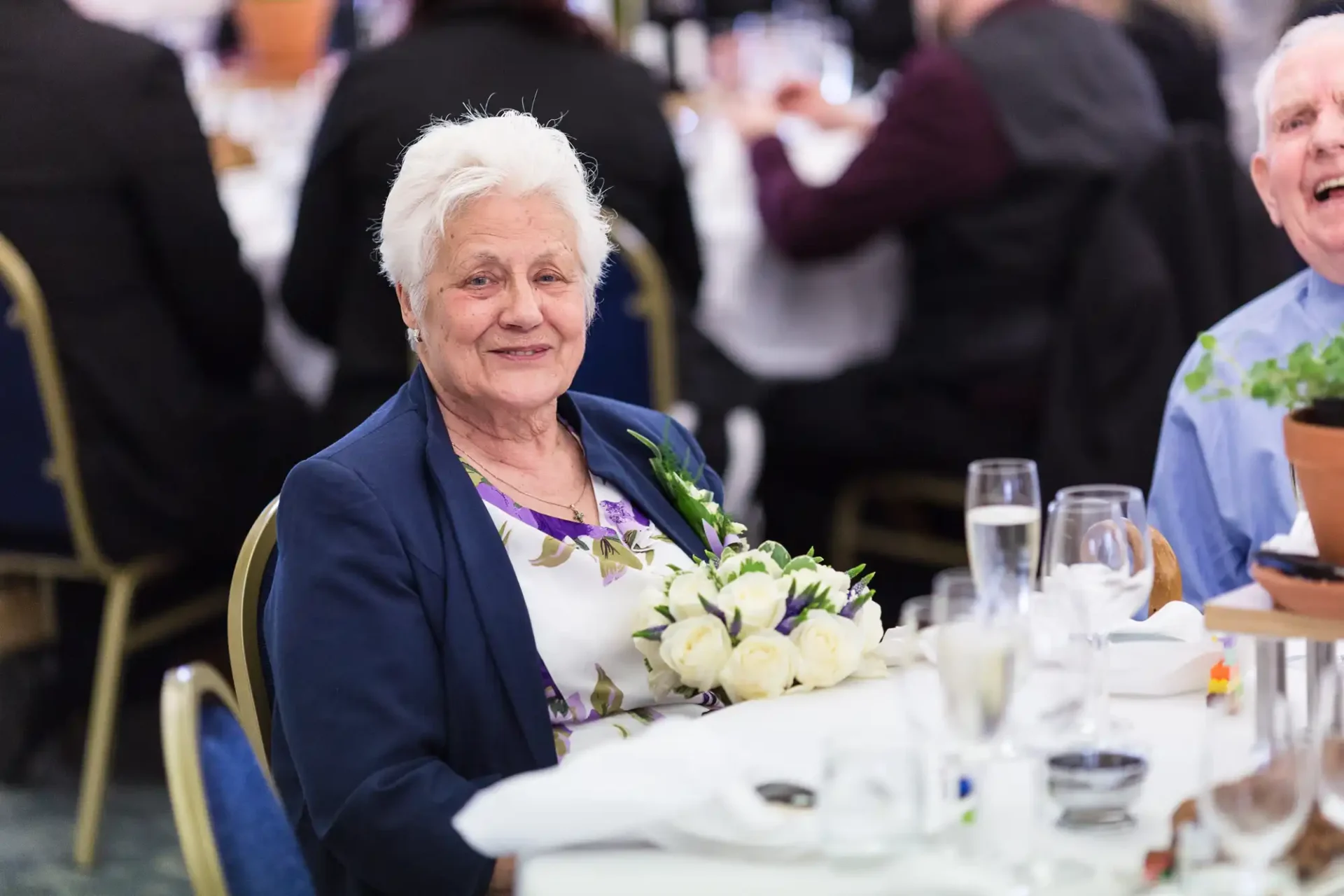 Elderly woman smiling at a banquet, holding a bouquet, with a man laughing in background.